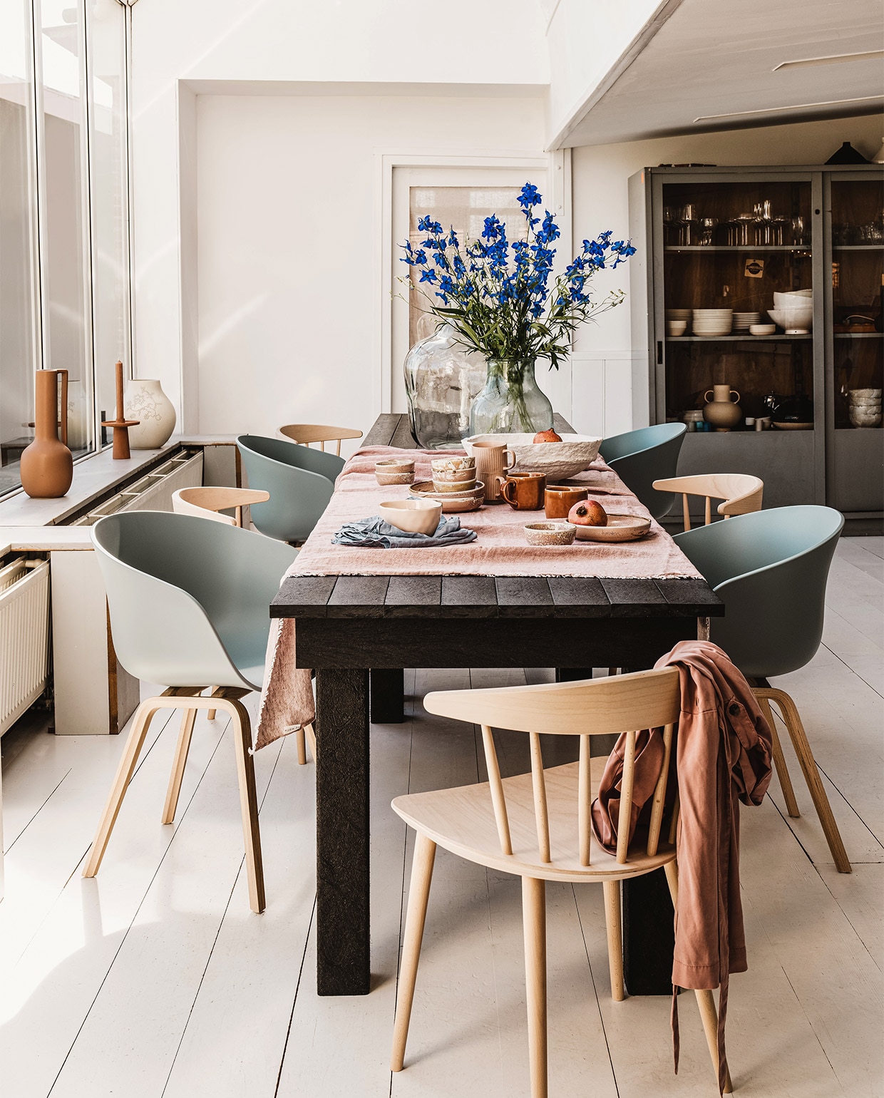 Tafels Standard Long Tables, gerecycled plastic en staal, ook geschikt voor buiten, 60x180x76 cm (bxlxh) per stuk € 1876,- (inekehans.com). Lichtblauwe eetkamerstoelen About a Chair AAC22 van Hay, per stuk € 279,-, beige houten eetkamerstoelen J104 van Hay, per stuk € 255,- (vtwonen.nl). Flesvaas Recycled, 56 cm (h) € 49,95, vaas Evita transparant large € 29,95, witte papier-maché schaal € 49,95, terra mok Svelte € 12,95, roze kom Good Morning € 13,95 (loods5.nl). Roze schaaltje Home Chefs Ceramics € 7,55, roze diep bord Home Chefs Ceramics € 15,95 (hkliving.nl). Roze linnen tafelkleed in kleur Blush, 145×270 cm € 149,-(bymolle.com). Lichtblauwe linnen servetten van H&M Home, set van 2 € 9,99 (hm.com). Roze blouse € 29,95 (zara.com). Terra kandelaar op vensterbank, vanaf € 5,99 (karwei.nl). Overig privébezit. JA | vtwonen 06-2022 | styling Moniek Visser | fotografie Denise Pronk