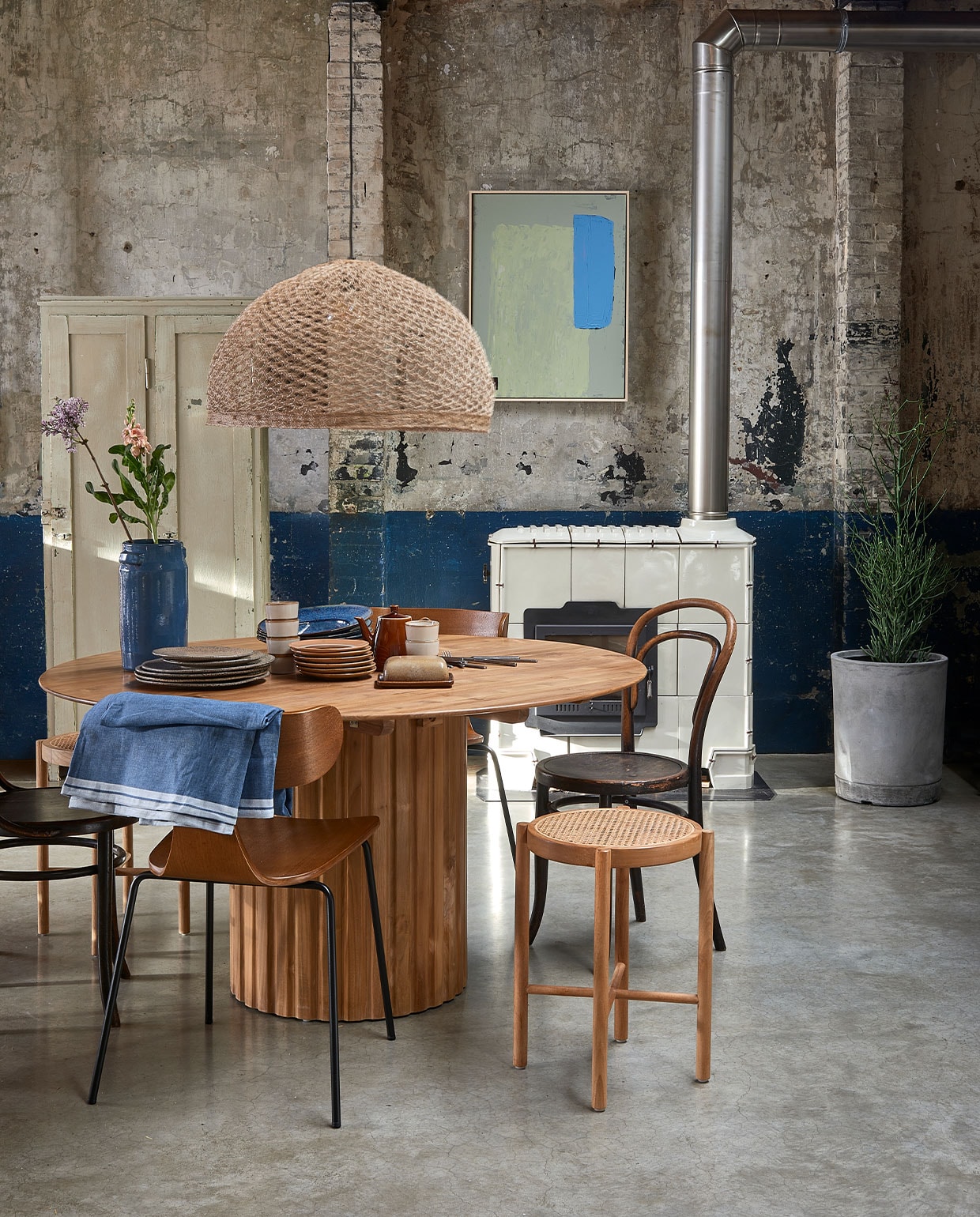 Teakhouten eettafel Pillar, 140 cm (Ø) € 1600,-, retro webbing kruk € 149,-, diverse borden vanaf € 7,50, mok € 8,50, abstract schilderij, 60×80 cm € 349,- (hkliving). Hanglamp, handgeweven uit India, 65 cm (Ø) vanaf € 95,-, betonnen plantenbak XL, 40×45 cm (Øxh) € 195,- (boxworx). Thonet stoel € 195,- (neeflouis.nl). Houten stoel Form met metalen poten € 199,- (deeekhoorn.com). Houten lockerkast € 275,- (vandijkenko). Tafelkleed 220×145 cm € 17,95 (ikea). Blauwe vaas € 53,- (fonq.). JA | vtwonen 03-2022 | Styling Fietje Bruijn | Fotografie Dennis Brandsma\n