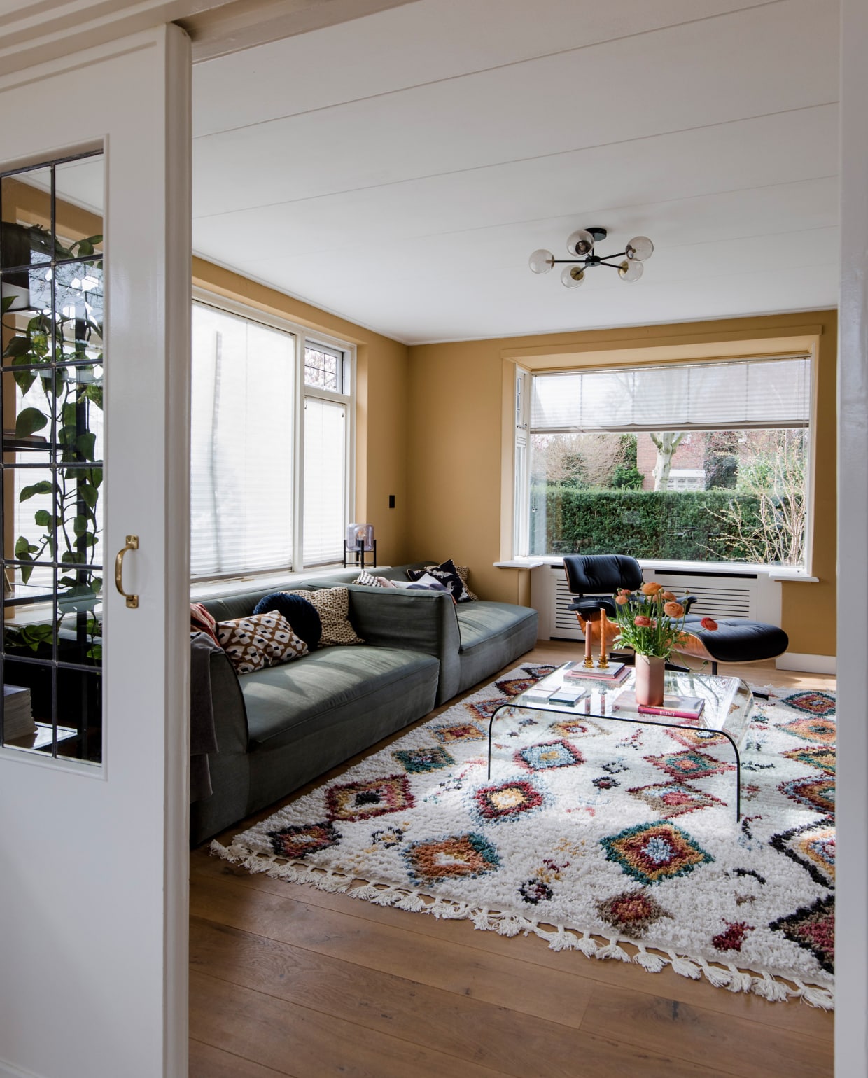 Glazen salontafel Fiam, vloerkleed Sezze van Trendcarpet, plafondlamp Globe van Made, Eames Lounge Chair in palissander van Vitra, vaas uit kringloopwinkel, kussens van Karwei, HKliving en Dutch Decor, kandelaars Kwantum, vloer wit geolied eikenhouten parket via Anderz, radiatorbekleding Cando, raambekleding Verasol. Muurverf Ginger Gold Le Noir et Blanc, overig onbekend. vtwonen 12-2022 | fotografie Corine Hoge Bavel