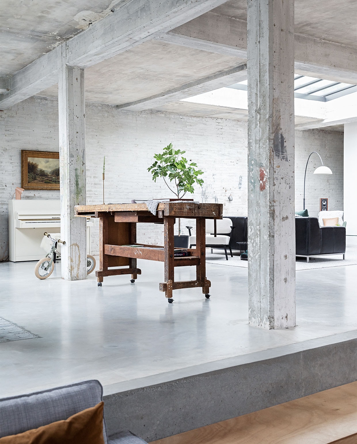 Vloer gevlinderd beton. Hardstenen waaltjes Spotted Bluestone. Werkbank erfstuk meubelfabriek. Kinderfiets Trybike. Witte fauteuil Ode Lounge Chair van Studio Henk. Bank Ikea. Booglamp Venled. Piano en schilderij erfstukken. Lamp op piano Elleke Rutten. JA | vtwonen 01-2022 | styling Ilona Jongepier  | fotografie Dana van Leeuwen