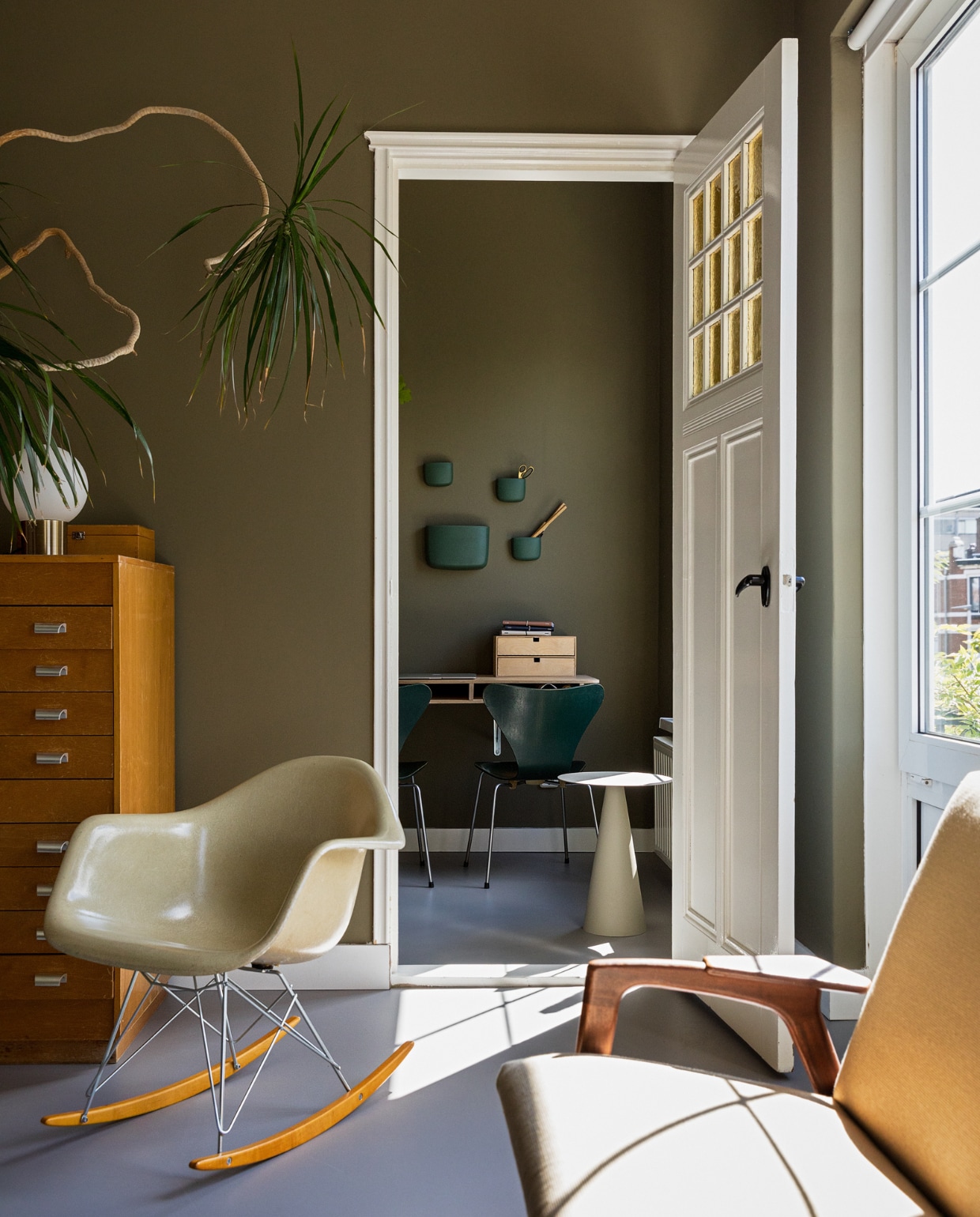 Muurkleur RAL 7002. Vintage plastic schommelstoel RAR van Eames voor Vitra. Bank van Yngve Ekstrm voor Pastoe. Oude papierkast uit een school. Vlinderstoel van Fritz Hansen via Vintagemarket12. Bureau zelfgemaakt. Bijzettafel Karwei. Kistje en bollamp rommelmarkt. Wandbakjes Normann Copenhagen. Overig onbekend. vtwonen 10-2022 | Styling Ilona Jongepier | Fotografie Dana van Leeuwen