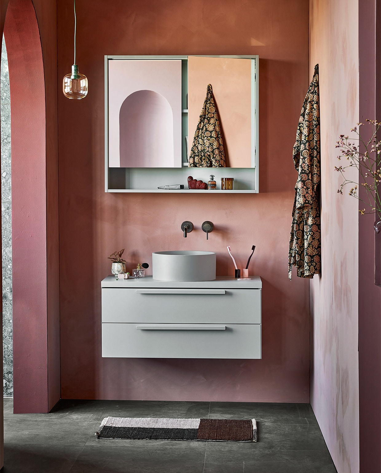 Wastafel uit vtwonen collectie. M.m.v. vestingh.nl (kleur op de muur Terra Pink No.102, kleur van kolom en rand toog Merlot Mania No. 123), zarahome.com (sloffen), vtwonen.nl (hanglamp Amp van Normann Copenhagen), hay.nl (bruine glazen opbergpot met deksel Container S, vloerkleed Burgundy, tandenborstel Tann, sculpture W&S Moguls Burgundy), nordal.eu (gekleurde glazen kandelaars, roze vaas, gespikkeld drinkglas, roze marmeren bakje), stories.com (parfum, tubes, sieraden). Overig privébezit. vtwonen 05-2022 | productie Tanja Saarberg | styling Kim van Rossenberg | fotografie Peggy Janssen