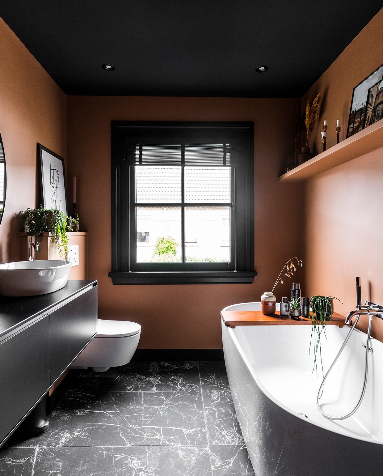 Badkamer met wandkleur Tasty Toffee Flexa, marmer look vloertegels Caesar Anima Nero mat Intercodam, bad Luca Sanitair, kraan Hotbath, badplank zelfgemaakt en in teakkleur gebeitst. Op de houten plank foto’s en souvenirs van vakantiereizen. Kozijn en raampartij zijn een renovatieingreep bedoeld voor extra licht. NEE | vtwonen binnenkijken special 2022 | producie CoCo Features | fotografie Muk van Lil, Chiela van Meerwijk