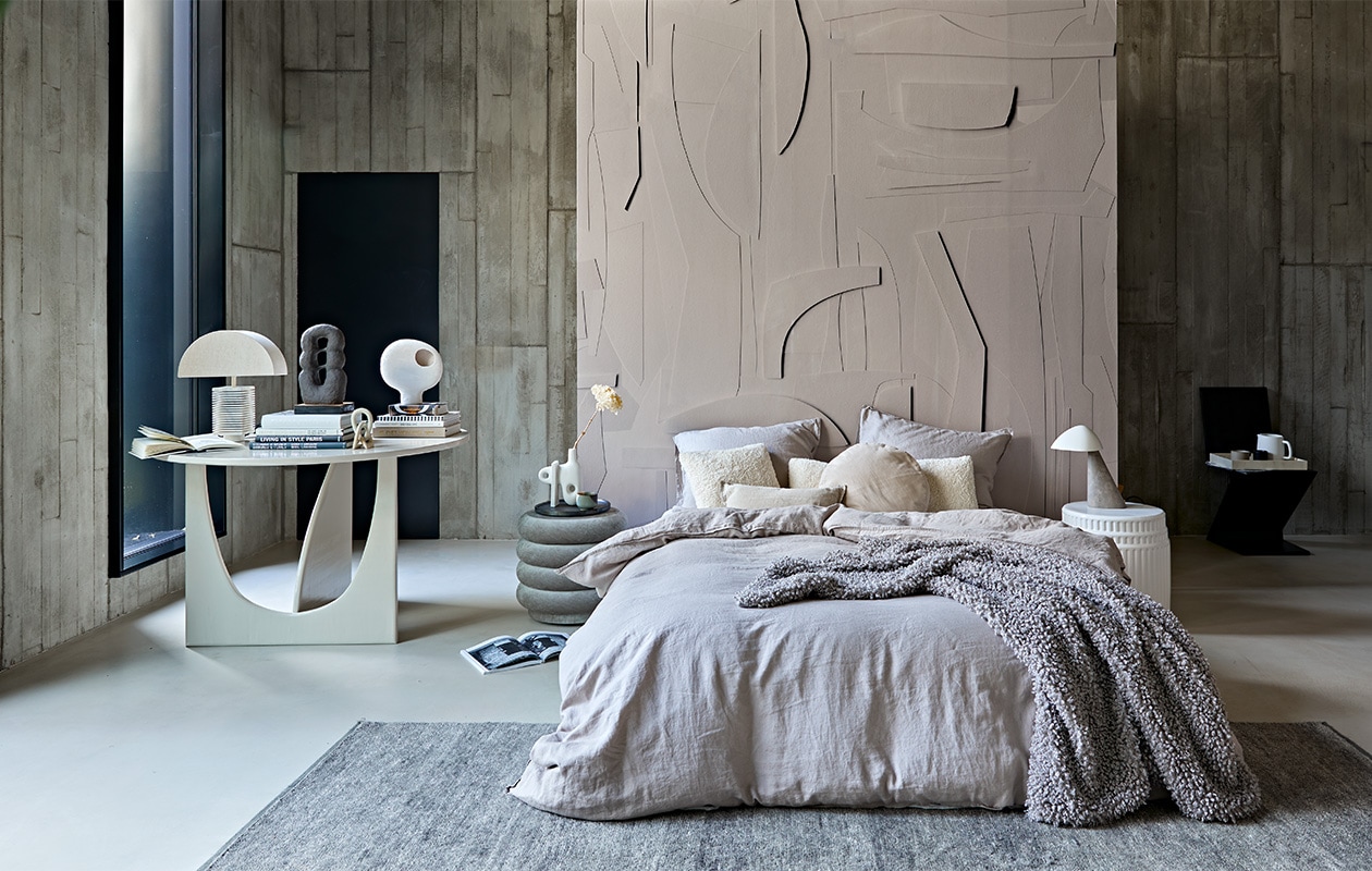 Behang op roomdivider White Spirit, op maat geprint, prijs op aanvraag (pascale-risbourg.com). Taupe-kleurig linnen beddengoed, 220×240 cm € 99,-, plaid van imitatie schapenvacht, 110×150 cm € 69,99 (hm.com). Grijze poef van Ferm Living, 42×40 cm (Øxh) € 679,- (edha-interieur.nl). Keramieken bijzettafel Kolos van Mini Forms, 45×46 cm (Øxh) € 1325,-, vloerkleed Lavo van Kvadrat, 300×200 cm € 2208,-(kasstoor.nl). Zwarte vintage zigzagstoel van Rietveld, prijs op aanvraag (vintagehoarder.com). Tafel 120×75 cm (Øxh) vanaf € 750,- (com-anders.nl). Vaas met gat op de stapel boeken € 39,95, teddy kussen 30×80 cm € 49,95, teddy kussen 40×40 cm € 29,95 (urbannatureculture.nl). Bruine sculptuur Guaja van klei, 18x8x33 cm (bxdxh) € 450,-, kleine stenen sculptuur Kisii Stone op tafel € 25,- (studio-yen.com). Links op grote tafel tafellamp Hexagon L, kap € 72,95, marmeren voet € 149,95 (hkliving.nl). Sculpturale vaas van ontwerper Jonathan Adler naast bed, € 209,- (ankevangoor.com). Klein linnen kussen, 30×50 cm € 15,99 (zarahome.com). Dienblad vanaf € 69,- (luxurybynature.nl). Keramieken waterkan van ontwerper Vincent van Duysen van Serax € 25,- (vtwonen.nl). Tafellamp Fellow met marmeren voet rechts naast het bed van Fredericia € 864,- (designtales.nl). Wandbekleding van locatie. Overig privébezit. JA | vtwonen 02-2021 | styling Cleo Scheulderman | fotografie Jeroen van der Spek