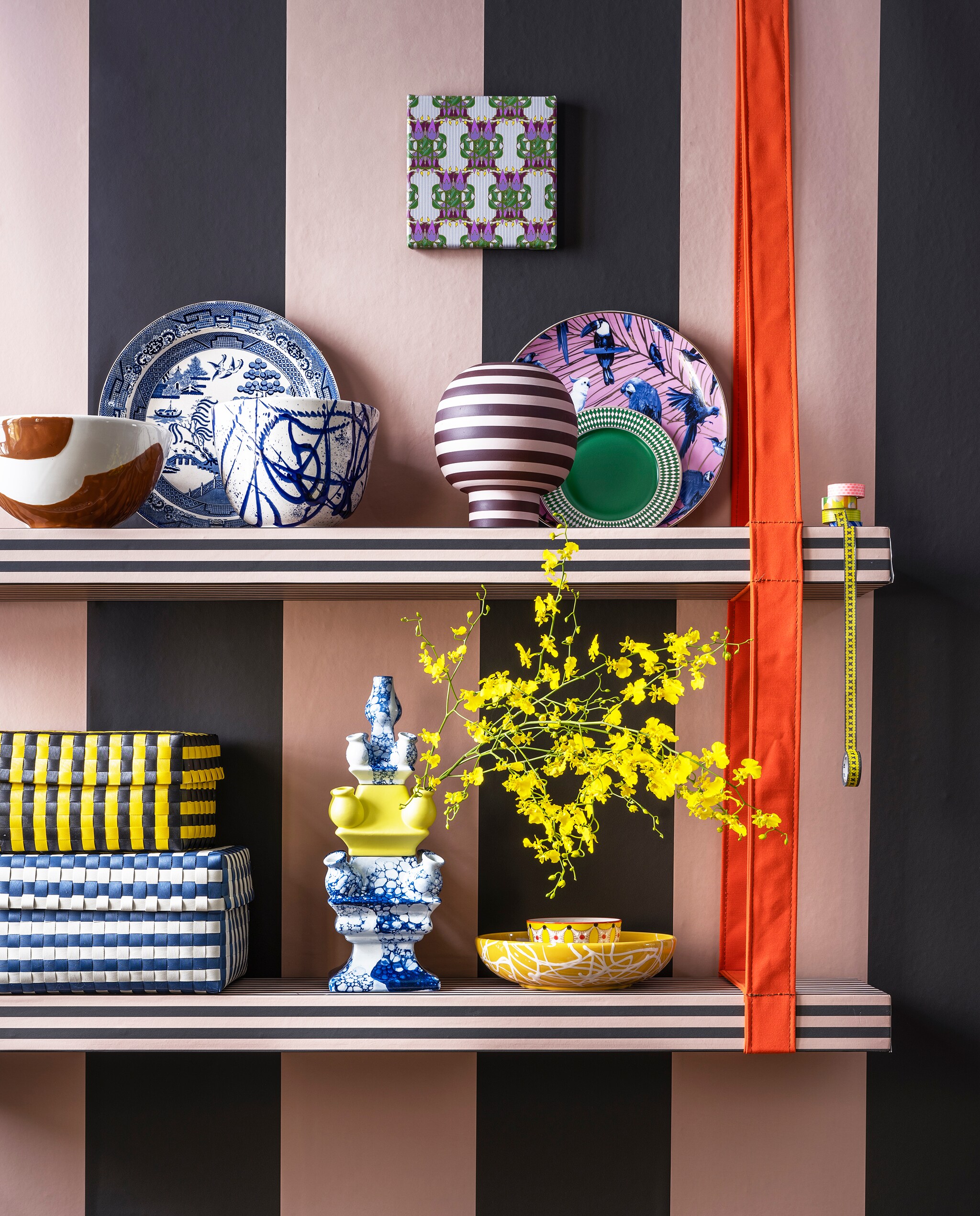Planken met ophangsysteem van oranje band van Combitex, prijs op aanvraag (com-anders.nl). Roze-groen streepbehang Thick Lines van Ferm Living, per rol € 79,- (misterdesign). Onderste plank: opbergdozen van gevlochten kunststof vanaf € 49,- (hay), 3-delige vaas € 236,- (heinendelftsblauw), gele schaal met spetterdessin € 49,95 (homata.fr), kommen van Pols Potten, per 4 € 53,80 (vtwonen.be). Bovenste plank: terra en witte schaal € 49,95 en blauw-witte schaal met spetterdessin € 49,95 (homata.fr), vintage bord tegen muur € 25,- (hetgroteavontuur.nl), gestreepte vaas van Aytm € 135,- (goeds), groen ontbijtbord Chess, per 4 € 82,50 (polspotten), bord met vogels inclusief ophangsysteem € 39,95 (heinendelftsblauw), Japanse tape vanaf € 3,- (ikigai-designbrands.com). Kunst op canvas Tulpen art deco van Iyaan, 20×20 cm € 49,- (werkaandemuur). vtwonen 06-2022 | styling Marianne Luning | fotografie Stan Koolen