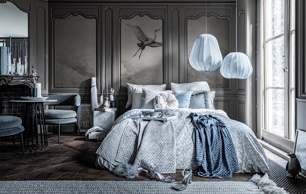 Behang op achterpaneel (wallanddeco.com). Kandelaar met takvorm van glas, witte vaas op tafel (frozenfountain.nl). Witte marmeren kandelaar met boog, blauwe en grijze kaarsen (loods5.nl). Marmeren tafeltje, wit vierkant nachtkastje, blauwe vaas,, (badenbaden.nl). Grijze stoel (edha-interieur.nl). Witte schaal op tafel groot, kleine kandelaar op schouw rechts, vaasje op nachtkastje, vloerkleed (sukha.nl). Blauwe vaas op tafel van Hay, grijze bollamp van Bolia groot, klein, zilveren pinguïn, grijze glazen , kan van Ferm Living, transparant glas naast bed van Ferm Living (vtwonen.nl). Zilveren hakken, boek op nachtkastje Noma, donkergrijs boek op de grond Christian Dior, wit linnen dekbedovertrek, rond kussen op bed, zilveren doosje op nachtkastje (hm.com). Blauw-wit dekbedovertrek met print, zilveren plaid, bijpassend kussen, sloffen met vachtje, grijs kussen, blauw kussen (zarahome.com). Doosje links op boek, boekjes op doosje (letoile.store). Blauwe ochtendjas, wit dienblad op bed, grijs-witte mokken op bed, witte pen in vorm van veer (maisonnl.com). Kunst in lijst op de grond (tinystories.nl). Beige stoffen vogel (homestede.nl). JA | vtwonen 02-2020 | styling Liza Wassenaar | fotografie Sjoerd Eickmans