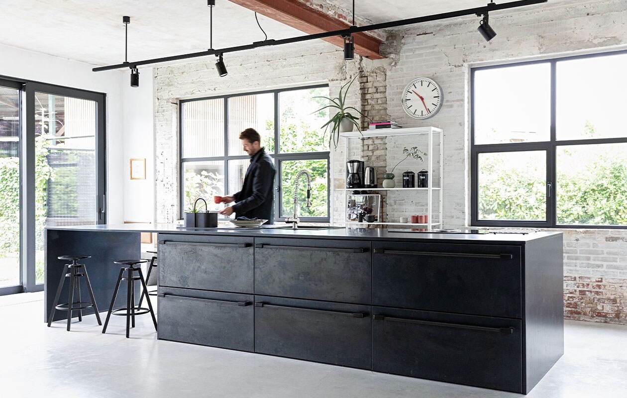 De keuken is een eigen ontwerp, binnenwerk op maat gemaakt door Marcel Hermans, beplating en werkblad van blauwstaal Schouren Metaal. Railarmatuur Venled. Toolbox Ferm Living. Krukken en open kast Ikea. Klok onbekend. Keukenblok eigen ontwerp, maatwerk, afgewerkt met geglazuurde tegels Praxis. Houten wand Okoume platen Bouwcentrum Driessen. Houten schappen koffiebar resthout meubelfabriek, deels geverfd in kleur Geordend van Histor. vtwonen 01-2022 | styling Ilona Jongepier | fotografie Dana van Leeuwen