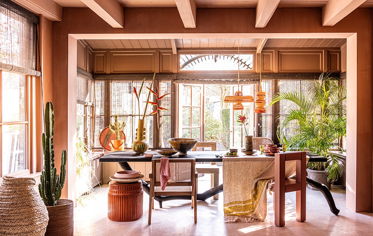Verfkleur op wand Truffel van Carte Colori, 2,5 liter, rieten stoel rechts achter tafel, leren wijnkoeler in raam, plafondlamp Maron, prijs op aanvraag, donkerbruine raambekleding Indivipro (brechtmurreatelier.nl). Zwarte tafel, 3 meter (l) (wdstck.eu). Beige beklede stoel, dezelfde stoel roze geschilderd (com-anders.nl). Oranje keramieken tafel, 58×35 cm (kasstoor.nl). Bloem in vaas en cactus (plantje.nl). Grote rieten mand, grote groene vaas, hoge groene schaal, kleine groene schaaltjes, per stuk, kleine groene vaas op tafel (householdhardware.nl). Rieten schaal in raam, mand links onderdeel salontafel, lichte rieten schaal op kruk, houten stoel met rotan, tafellaken met gele streep, 110×180 cm, groen wijnglas, linnen servet over stoel, 45×45 cm, hanglamp links, hanglamp rechts (couleurlocale.eu). Houten lepel op roze bakje (koconceptstore.nl). Rode kom Merci van Serax, 15 cm (Ø) (vtwonen.nl). Roze schaal met ore, langwerpig roze bakje, roze bord, oranje bord (sissy-boy.com). Groot bruin bord, bruine schaaltjes (letoile.store). Bloempot privébezit. vtwonen 07-2021 | styling Liza Wassenaar | fotografie Sjoerd Eickmans