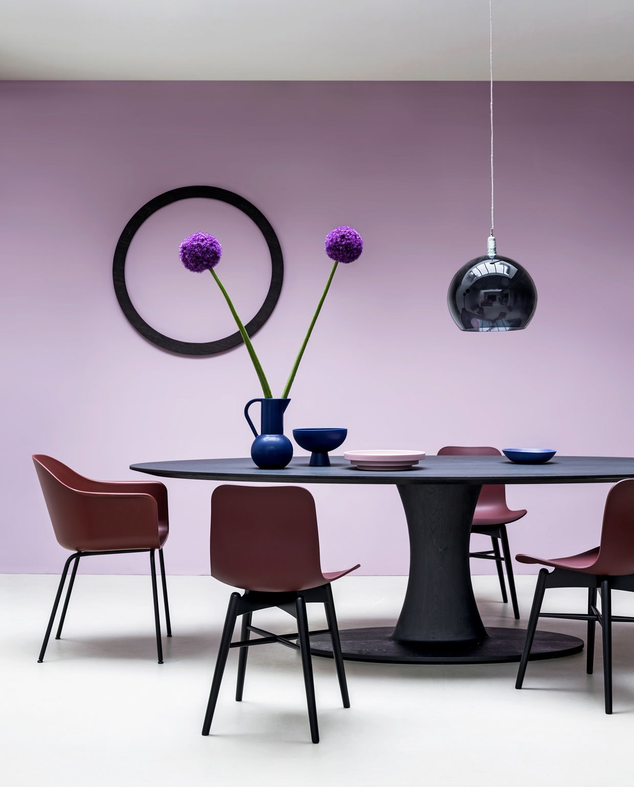Muurverf Washed Lilac van vt wonen (karwei.nl). Tafel van massief eiken in bijna zwart, 260x120x76 cm (lxbxh) (casagitane.com). Stoelen van Norr 11 (eijerkamp.nl). Stoel Harbour met armleuning van Menu, glazen lamp van Ebb &amp; Flow, Ø 39 cm (goeds.nl). Kan met oor, schaal op voet (groenenakker.nl). Roze schaal in Memphis stijl, soepbord (hkliving.nl). Houten cirkel privébezit. vtwonen 10-2019 | styling Marianne Luning | fotografie Stan Koolen