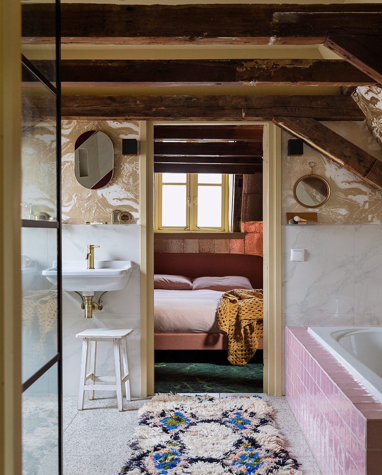 Terrazzo vloertegels, keramische wandtegels met marmerprint, kraan van geborsteld koper, roze badtegels en witte kruk herkomst onbekend. Behang Marble Gold 188 van Ferm Living. Wastafels Villeroy &amp; Boch. Ovalen spiegel &amp;Klevering. Ronde spiegel antiek. Kleed The Souks. In de slaapkamer: behang Antique Mirror in goud van Cole &amp; Son. Bed Made. Beddengoed Ikea. Sprei zelf gehaakt. JA | vtwonen 06-2021 | styling Ilona Jongepier | fotografie Dana van Leeuwen | tekst Renate Dahlmans