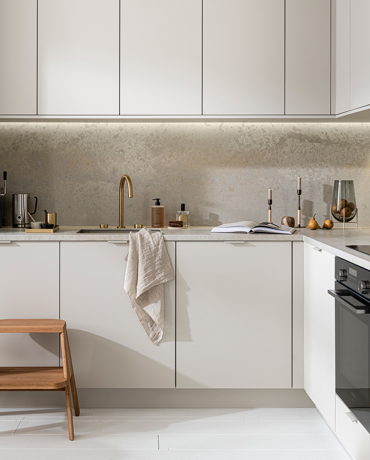 Keuken Basic Greige, vanaf € 21.500,-,  M.m.v. thefinestore.com (houten krukje van Hay, messing dienblad en beker van Ferm Living, bronzed brass kandelaren Duca van Menu), flinders.nl (vaas van gerookt glas op messing pootjes van Menu), kikubari.nl (spray van Lingua Planta), vtwonen.be (theedoek uit de vtwonen collectie), skins.nl (handzeep Suede Byredo), sjostrandcoffee.nl (melkschuimer). Overig privébezit. vtwonen 03-2021 | productie Tanja Saarberg | styling Kim van Rossenberg | fotografie Sjoerd Eickmans