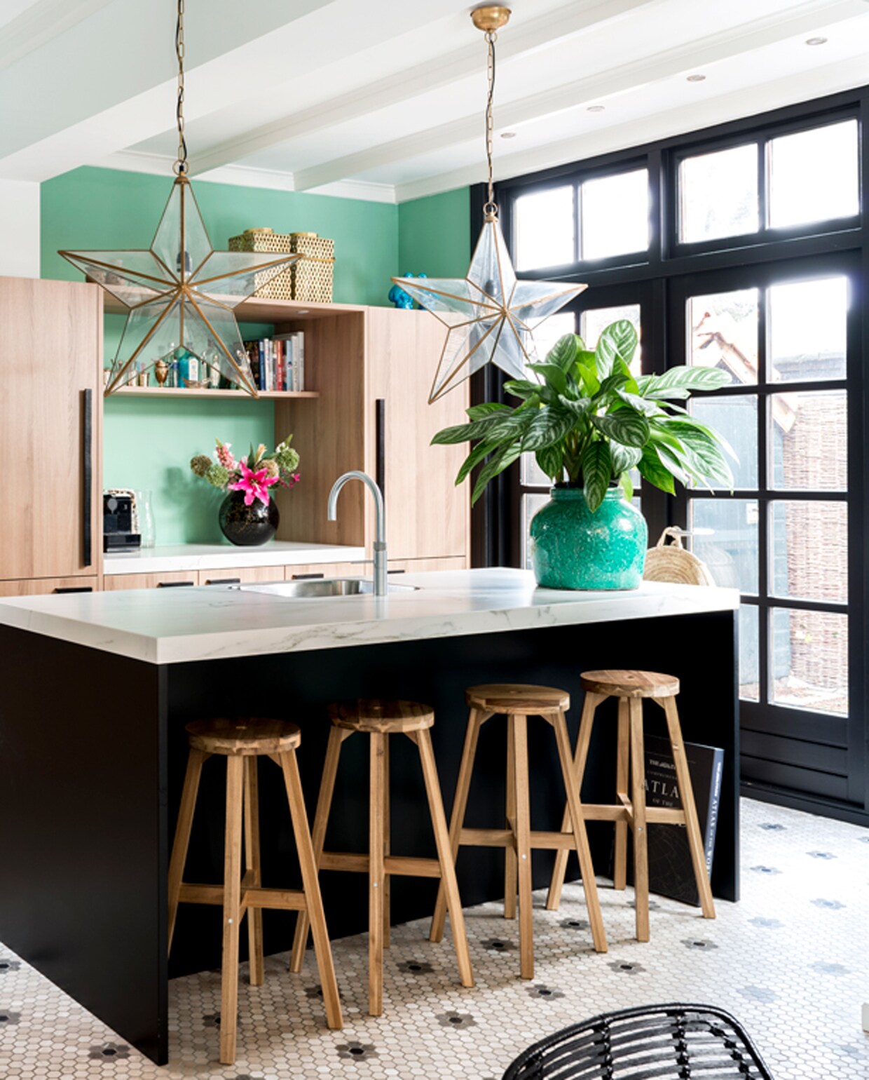 Keuken van Piet Boon. Stervormige lamp van Sissy-Boy. Plantenpot Mica Decorations. Vloertegels van designtegels.nl. Krukjes Ikea. Ananas links op het aanrecht komt uit Parijs, evenals de windlichten boven op de keukenkast, verkrijgbaar bij Studio Sophie. Muurverf kleur Arsenic van Farrow &amp; Ball.