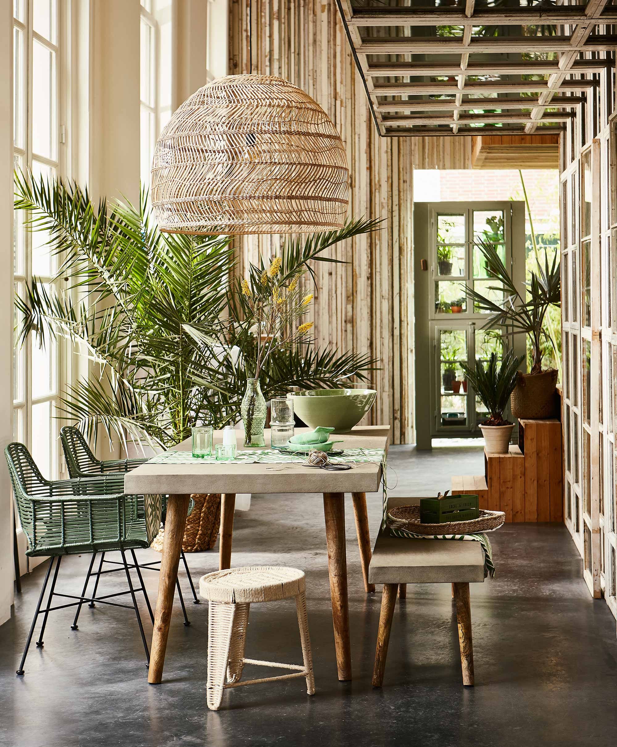 Tafel met betonnen blad en bijpassende bank (Nordal). Gevlochten hanglamp, groene rieten stoel (HK Living). Kruk van gevlochten touw, gevlochten schaal (Madam Stoltz). Lopers, groen met wit en groen kratje (H&amp;M Home). Karaf in de vorm van een vis, groene visvormige glazen schalen, lage visschaal van glas, laag groen glas, hoog groen glas, wit zoutvaatje, alles uit de collectie van Paola Navone (Serax). Glazen karaf met ribbel (Household Hardware). Grote groene aardewerken schaal (De Weldaad). Rieten mand met palm, gevlochten mand met palm op trap (Nordal). Groene kast (Combitex). Overig privébezit. vtwonen 07-2016 | Fotografie Jeroen van der Spek | Styling Cleo Scheulderman
