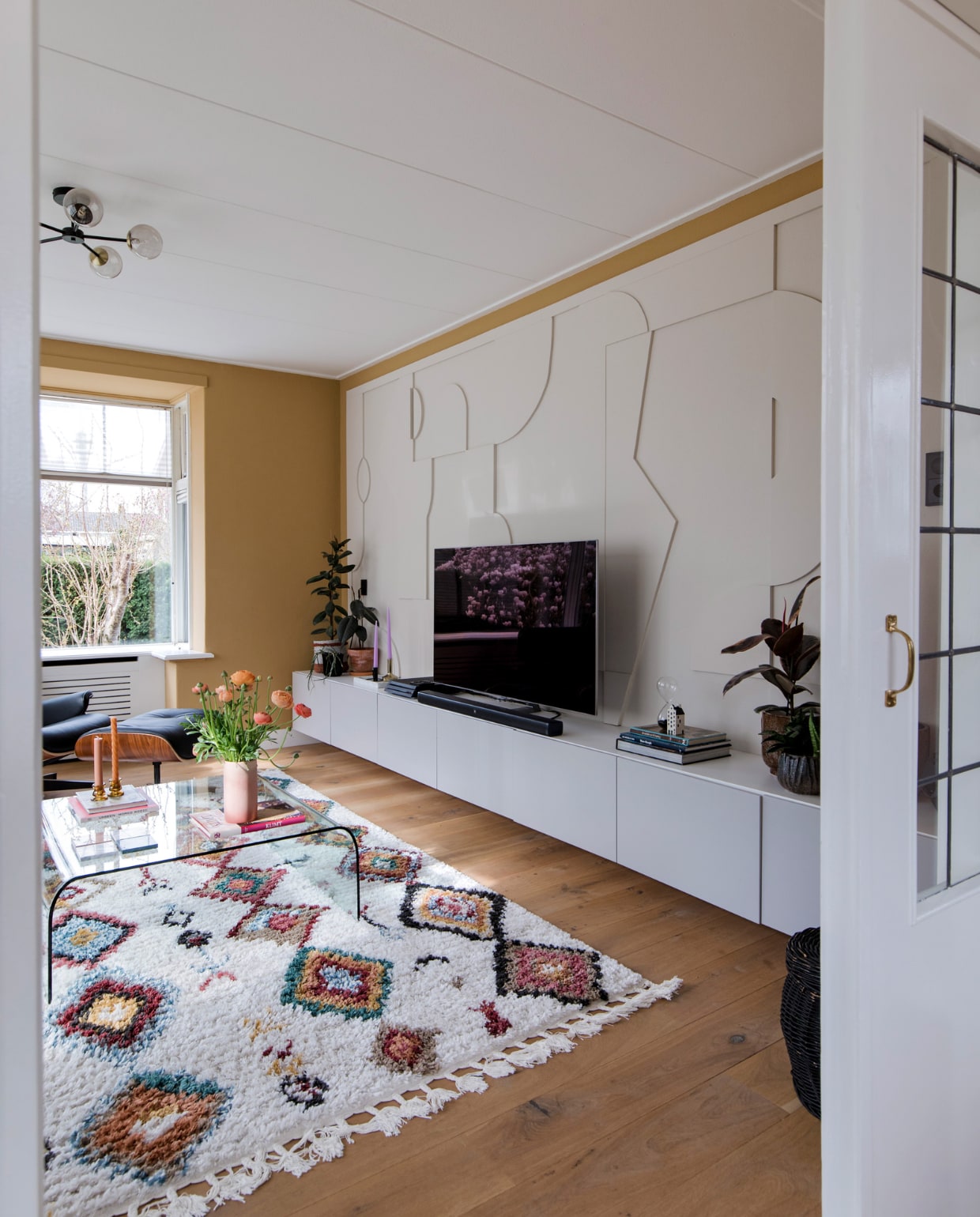 Salontafel Fiam, vloerkleed Sezze van Trendcarpet, Eames Lounge Chair en Ottoman van Vitra. Plafondlamp Globe van Made. Vaas kringloopwinkel, tv-meubel Ikea, 3D-wand zelfgemaakt, tv Samsung.Vloer wit geolied eikenhouten parket via &amp;Anderz, radiatorbekleding Cando, raambekleding Verasol. Muurverf Ginger Gold Le Noir et Blanc en Skimming Stone Farrow &amp; Ball.