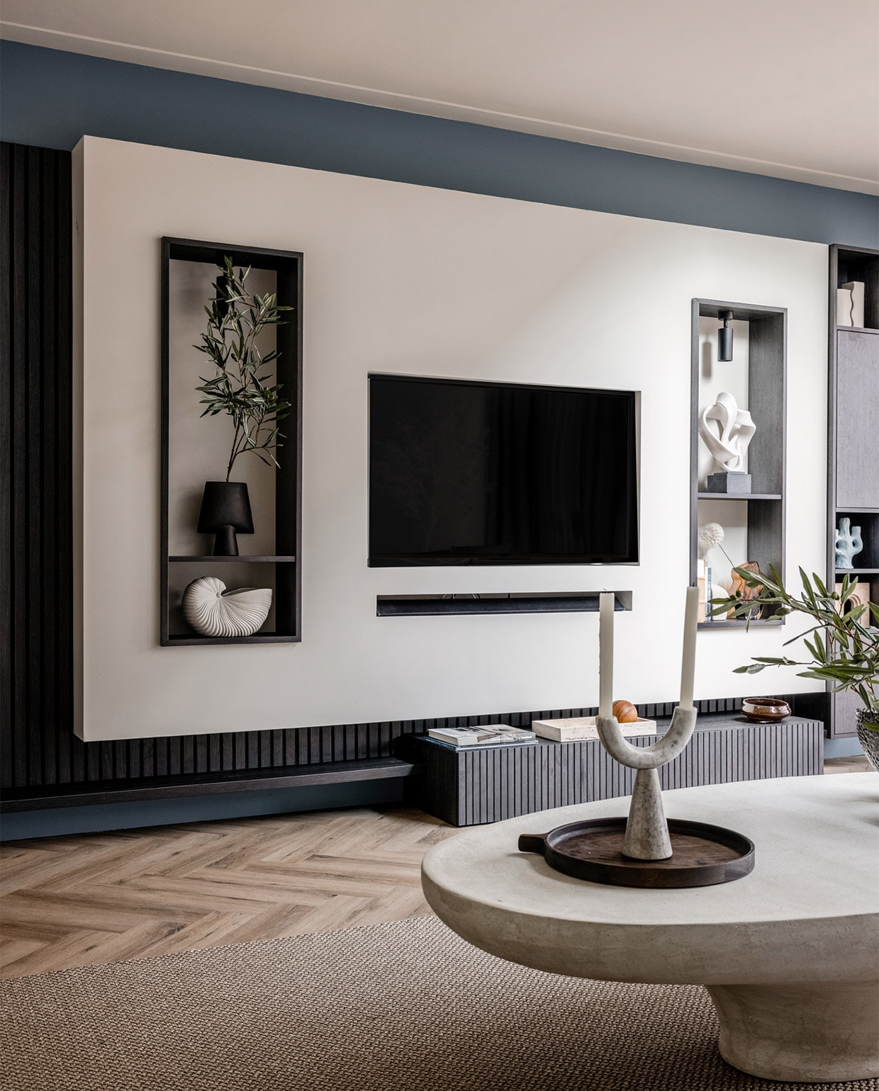 Flexa Creations muurverf Denim Drift (flexa). Plafondspots in de kast € 64,95 (lightinova.com). Wandmeubel met een verticale lattenwand en ledverlichting vanaf € 5800,- (meijermaatinterieurs). Sisal vloerkleed Luca in uitvoering 6704 € 2550,- (cunera.nl). Salontafel € 1599,- (lanena-home.com). Bank, per element vanaf € 850,-, donkergrijze vaas in de linker nis € 65,-, blauwe kandelaar € 38,50, zwart ornament op de salontafel € 22,99, vazen op de salontafel, per stuk vanaf € 185,-, houten dienblad € 169,- (eijerkamp). Witte schelpenvaas van Ferm Living € 85,15, blauwe plaid van Malagoon € 39,95 (vtwonen.be). Witte vazen in de kast rechts, set van 2 € 14,99, bruine vaas € 24,99 (hm.com). Notitieboek € 35,65 (badenbaden.nl). Beige object in de kast rechts € 249,-, dienblad waar de houten bal in zit € 169,- (stonedamsterdam.com). Marmeren siervorm op voet € 155,78, kandelaar op salontafel € 69,69 (nordal.dk). Kaarsen, per stuk € 1,- (hema). Olijftak 138217, prijs op aanvraag (silk-ka.com). Overig privébezit.