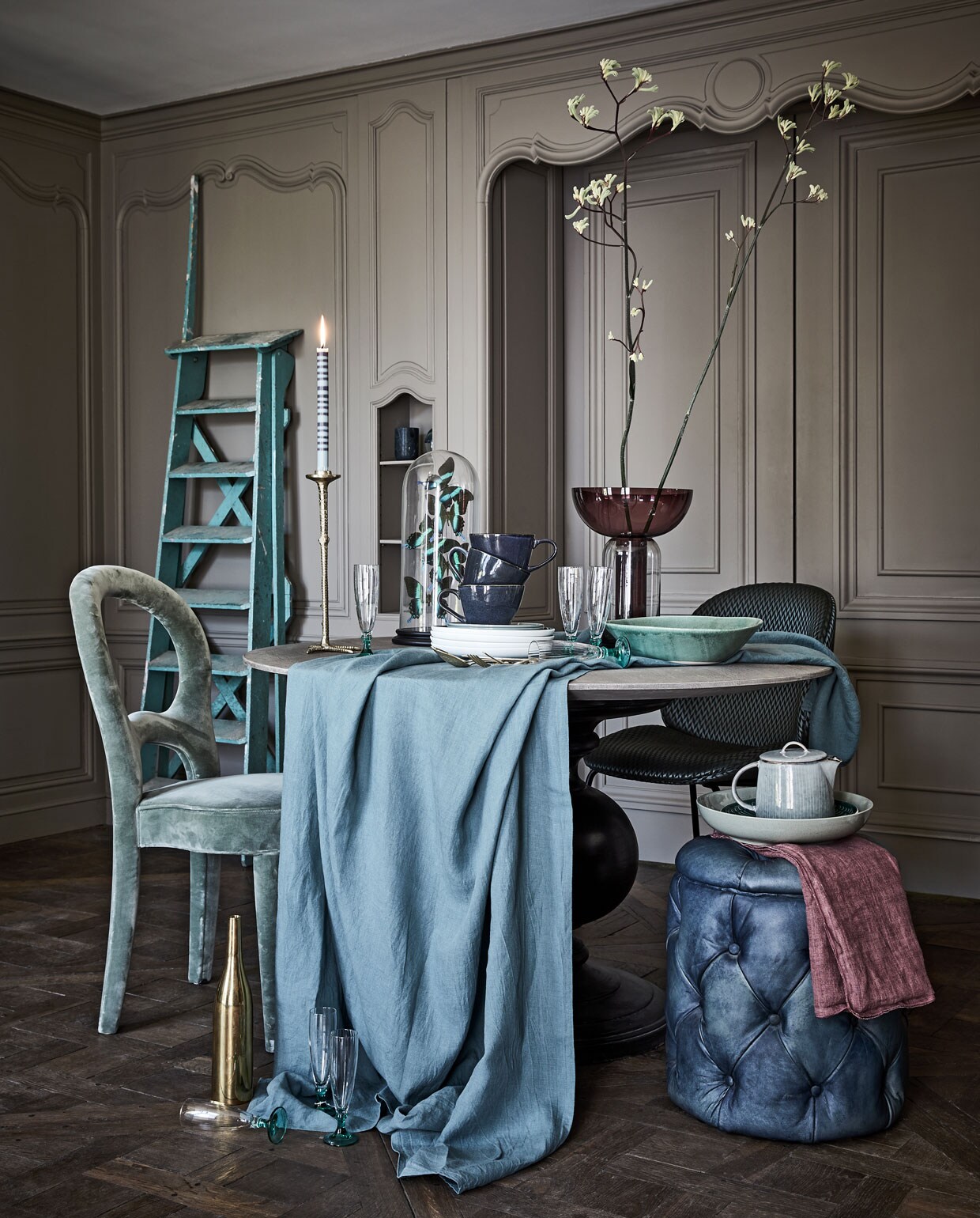 Tafel rond met zwarte poot Ø 130 cm, poef blauw (www.purefurniture.com). Stoel groen Promemoria (covanderhorst). Stoel midden, gouden fles (edha-interieur). Ladder (burbri.nl). Hoge vaas (homestede). Kaars, theepot (maisonnl.com). Borden It’s my match van Villeroy &amp; Boch, marmeren mok van Zuiver (vtwonen.be). Bestek goud vanaf (zarahome.com). Kandelaar, linnen tafelkleed, blauwe mokken (loods5.nl). Lichtblauwe schaal (hm.com). Glazen op groene voet (wildschut-antiques.com). Paars linnen (bungalow.dk). Stolp met kunstvlinder (weldaad.com). Groene schaal privébezit.