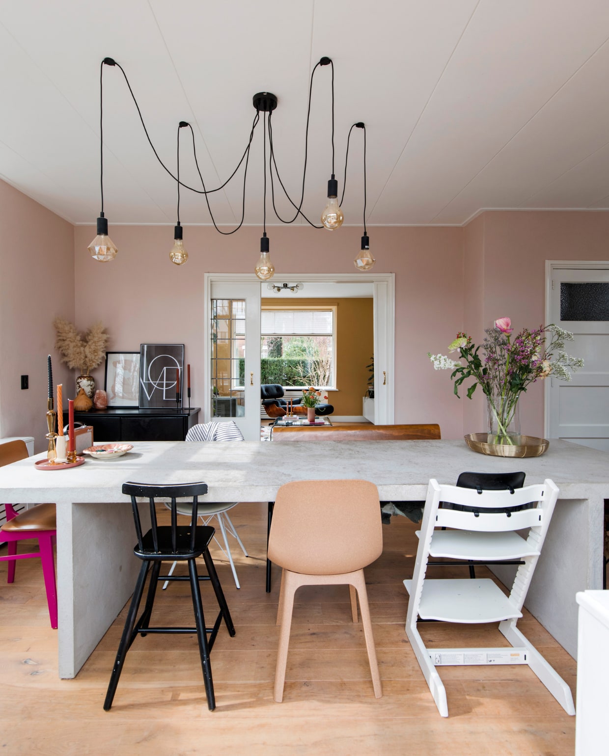 Tafel zelfgemaakt met beton ciré. Felroze stoel vintage. Zwarte kinderstoel, stoel Odger en lijsten Ikea. Witte kinderstoel Tripp Trapp van Stokke. Hanglamp Eglo. Eettafelbank vtwonen. Goudkleurige draadschaal Xenos, kandelaars Kwantum, vintage aardewerken schaal Flora Gouda, Chinese porseleinen vaas familiestuk, posters I am Fy. Muurverf Pink Nudity Flexa. Wit geolied eikenhouten parket &amp;Anderz. Overig onbekend.