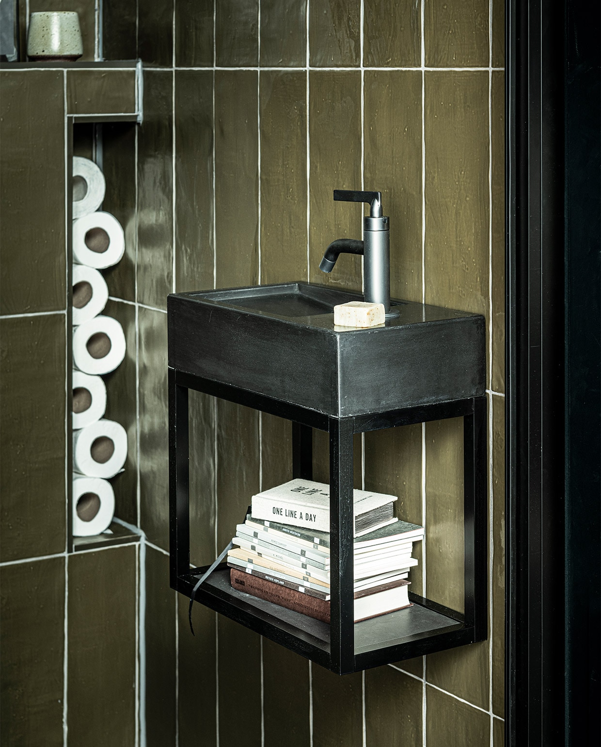 Fontein Tank in kleur Concrete Black, 40x22x10 cm (bxdxh) € 299,- (ook in Concrete en in White, hoogte 5,5 cm), metalen onderstel Fontein Frame in Zwart, 40x22x30 cm (bxdxh) € 339,-, hendel wastafelkraan Grip in kleur Black Steel € 459,- (ook in Charcoal, Bronze, Brushed en Chrome en met twee hendels), wandtegel Mediterranea in kleur mat Camouflage, 3,2×40 cm per m² € 61,75, vloertegel Classic Black mat, 74×74 cm per m² € 73,05 (ook in 74×148, 30×60 en 7,3×30 cm).WC uit vtwonen collectie: tegeldepot (toilet), ikea.nl (vaas), vtwonen.nl (fotolijsten).