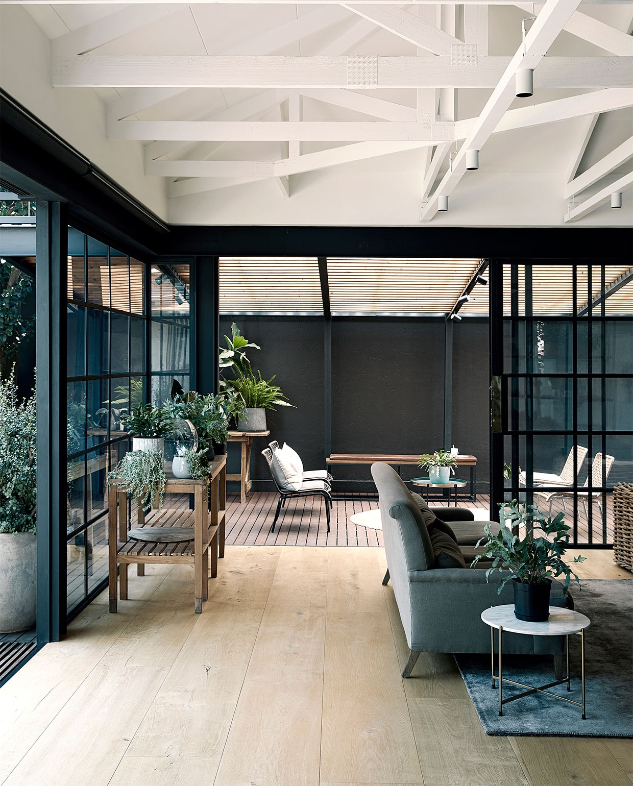 Bank Mezzanine Interiors. Rieten bank Weylandts. Bank rechts en stalen stellingkast Live Simple. Tapijt en ronde bijzettafel Hertex. Plantentafel en bloempotten kringloopwinkel. Overig onbekend.