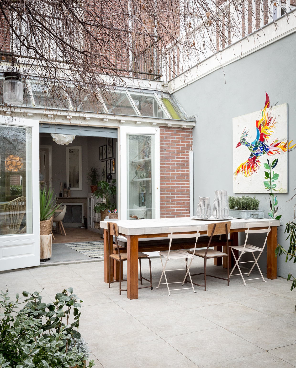 Muur buiten geverfd met buitenverf van de bouwmarkt in kleur gelijk aan vtwonen Ash. De eettafel (privébezit) is betegeld met tegels Neo Dessin Ambre uit de vtwonen-collectie by Douglas &amp; Jones, 20×20 cm per m² € 69,50, bestrating tegels Hormigon Grey uit de vtwonen-collectie by Douglas &amp; Jones, 60×60 cm per m² € 49,60 (www.vtwonen.be). Op de buitentafel: stalen bak € 20,- (www.neeflouis.nl). Rozemarijn per m² (www.greenlifestylestore.nl). Antiek dienblad aluminium small € 115,-(www.householdhardware.nl). Lantaarn Bars van House Doctor, 30 cm € 21,99, lantaarn Bars van House Doctor, 40 cm € 27,50 (www.vtwonen.be). Bistrostoelen in de kleur Linen € 64,- (www.extravert.nl). Oude houten vintage stoel (privébezit), soortgelijke stoelen € 69,- (www.ibizaoutdoor.nl). Kussen op de bank is op maat gemaakt door Meta Poulain. Lichtsnoer van House Doctor, 10-delig zwart € 94,95 (www.vtwonen.be). Overig privébezit.