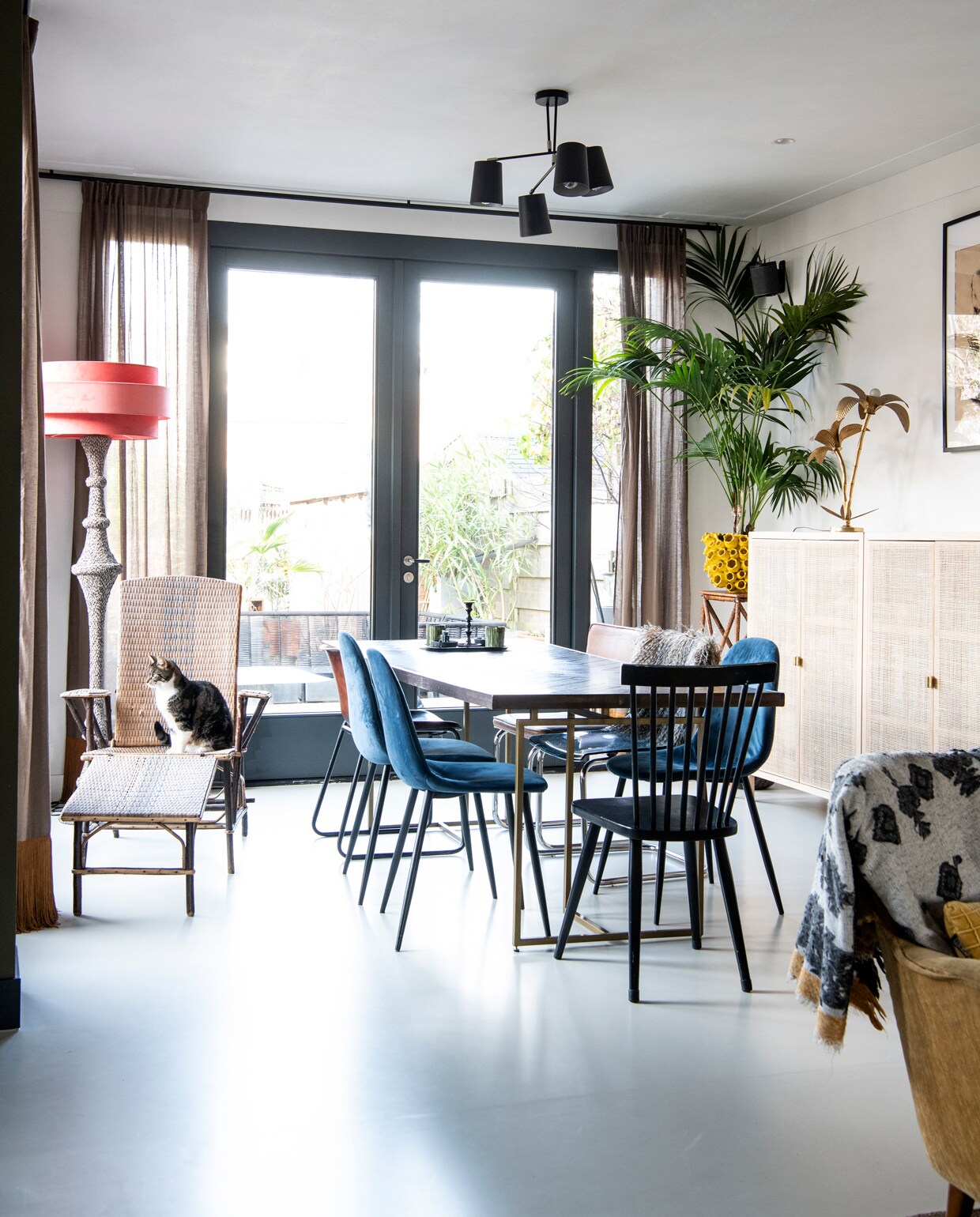 Vintage rotan loungestoel gekocht bij een garageverkoop. Lampvoet Loods 5, kap Madame Garage. Eettafel Dutch Bone. Blauwe stoelen Kwantum. Zwarte stoel vintage. Plafondlamp Rietveld Licht. Gele plantenpot Colmore Diga. Wandkastje met webbing Ikea. Overig onbekend.