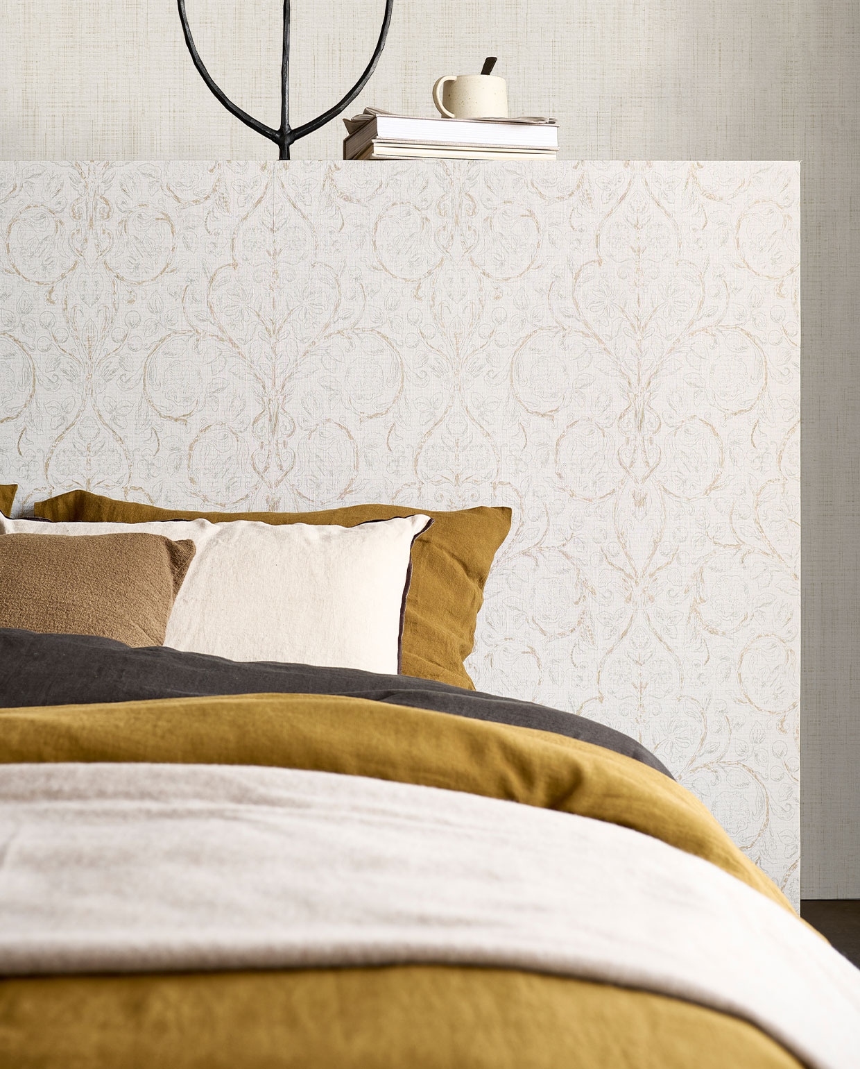 Behang op hoofdbord Classic Curl Light Grey, op achterste wand Linnen Gold, 53 cm breed, rol van 10 meter €29,99 (classic Curl ook verkrijgbaar in Sea Green).M.m.v. pure-original.nl (muurverf Fresco in kleur Old Flax), paviljoennoord.com (linnen dekbedovertrekken, linnen kussenslopen, olijfgroen kussen), letoile.store (mok), sissy-boy.com (metalen kandelaar, plaid), zarahome.com (wit kussen). Overig privébezit.