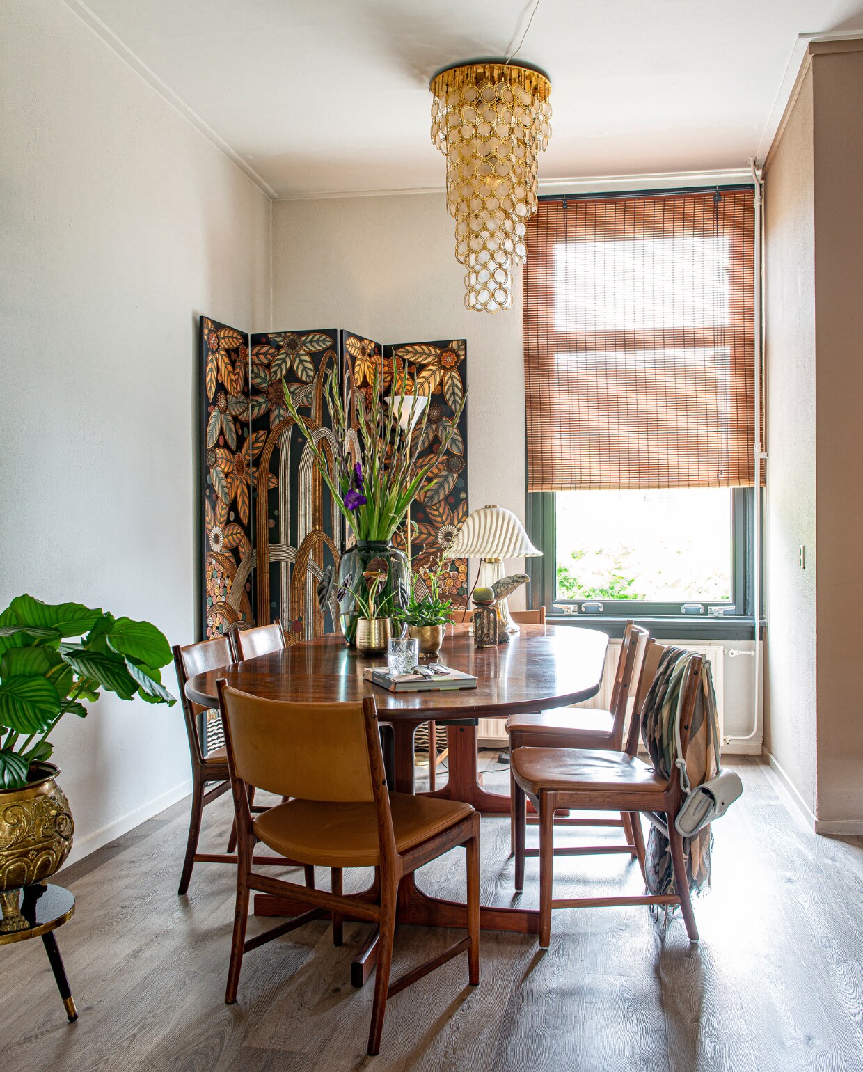 Lamp van Verner Panton, handgemaakt kamerscherm uit de jaren 80 in art-decostijl van Fournier Decoration, eettafel uit de jaren 50 met stoelen van Kai Lyngfeldt Larsen voor Søren Willadsen, stenen vogel op tafel herkomst onbekend, paddenstoellamp van Peill &amp; Putzler, Italiaanse staande lamp merk onbekend, vaas, bloempotten De Bloemenwinkel, oude koperen pot van een kringloopwinkel, plantentafel herkomst onbekend, sjaal van Humanoid, handtas van Heleen van de Veer.