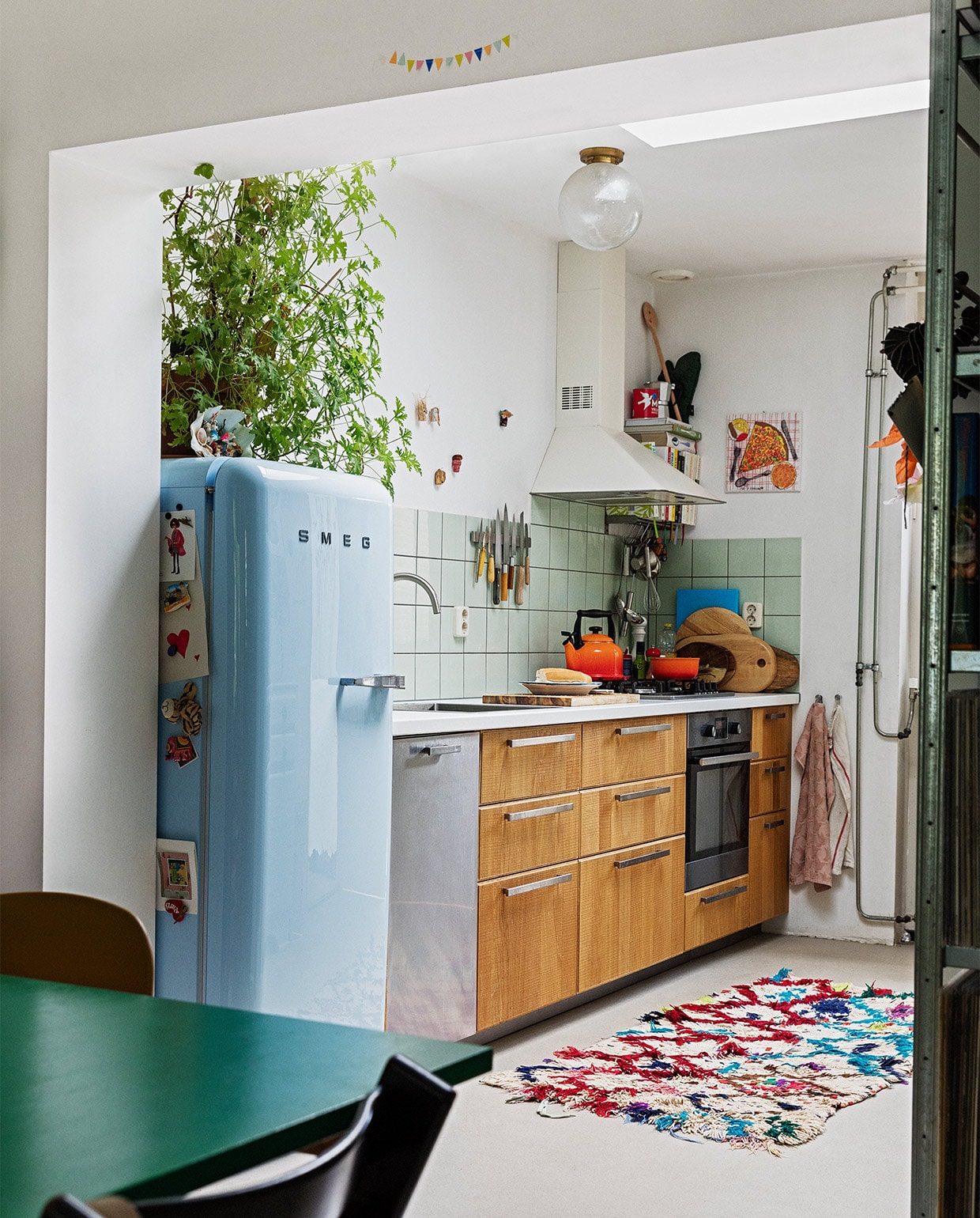 Koelkast Smeg. Geraniumplant zelf gestekt. Kitsch schelp op koelkast souvenir uit Griekenland. Magneet-souvenirs meegenomen van verschillende reizen. Fluitketel en pan Le Creuset. Messen Suncraft, Opinel, Dille &amp; Kamille, Pallarès Solsona. Snijplanken Hay, Ikea, Serax. Soeplepel Brabantia. Rvs keukengerei Dille &amp; Kamille. Schaaltje van een pottenbakkersmarkt. Afzuigkap en oven Ikea. Ovenhandschoen Kook. Pollepel Kookhuis aan de Maes. Handdoeken Sissy-Boy. Tapijt The Souks. Pizza-schilderij Oskar. Ronde plafondlamp lampenwinkel.nl.