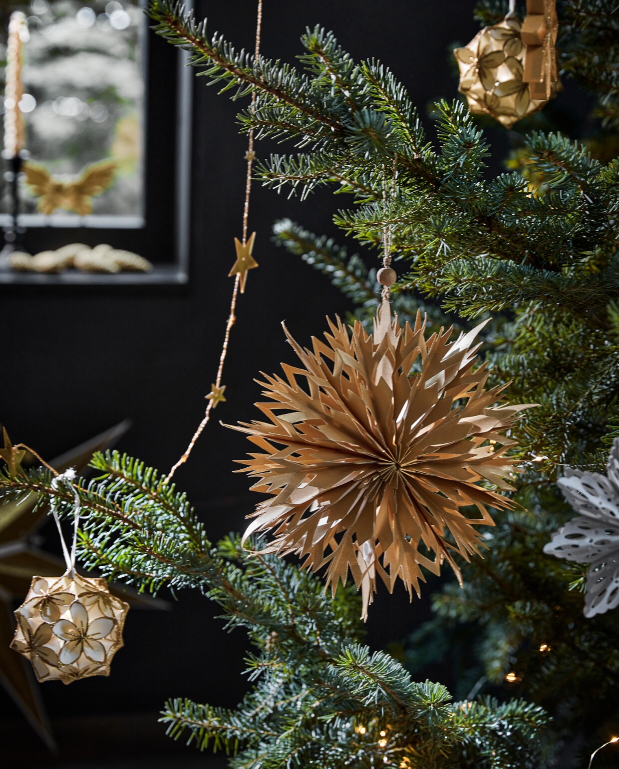 Goudkleurige metalen slinger met sterren, 210 cm (l) € 3,49, wit papieren ornament bloem met glitter, 10 cm (Ø) € 5,99 (ook verkrijgbaar in zwart met zilver). Goudkleurig papieren ornament, 20×5,5 cm (Øxd) € 3,29, goudkleurig ornament ster, 15×1,5 cm (Øxd) € 2,99.