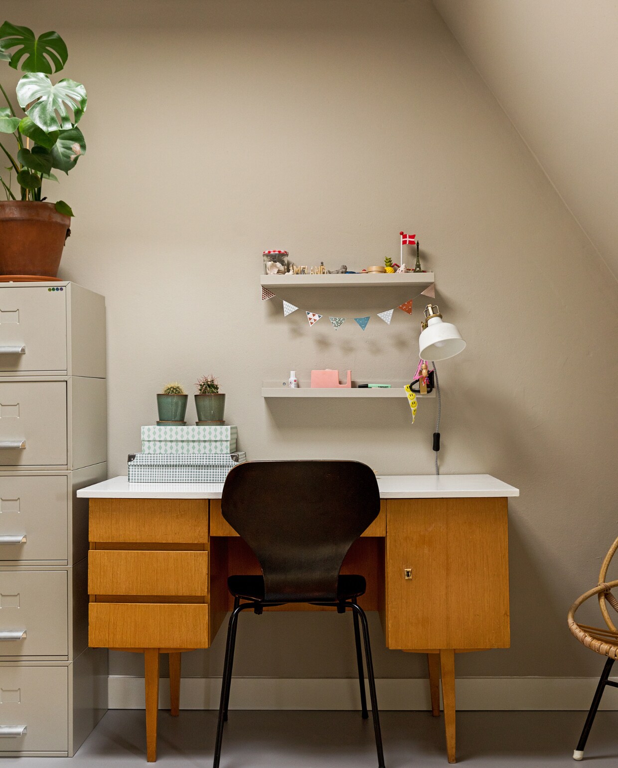 Muurkleur RAL 7044. Bureau, kast en rieten stoel Marktplaats. Stoel vintage. Vloerkleed, wandplank en lamp Ikea. Dozen, plant Nørd Conceptstore. Op de vloer ligt linoleum.