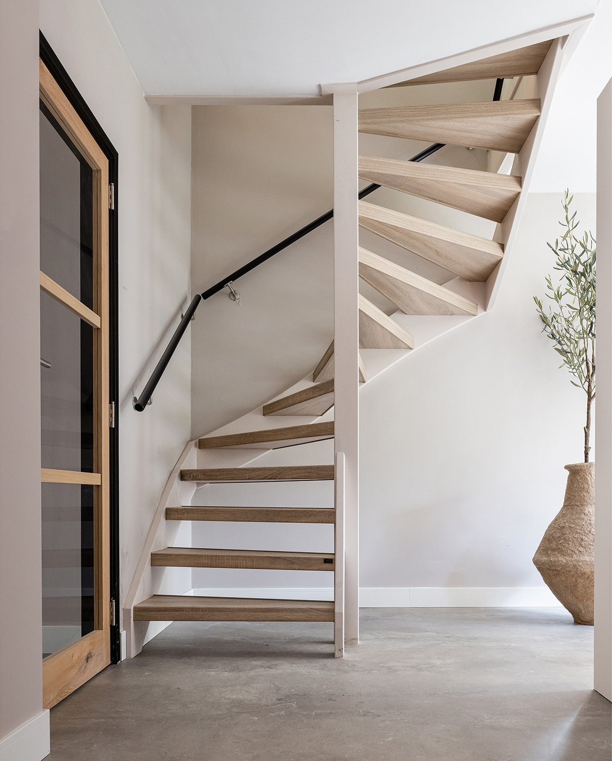 Pvc-vloer betonlook Dynamic Cemento 609 met plint, prijs op aanvraag (woninginrichting-aanhuis.nl). Trap bekleed met decor San Diego Oak, prijs op aanvraag, trapleuning rond, kleur Oak, prijs op aanvraag (upstairs.com). Vaas XL € 199,- (eijerkamp.nl). Olijfboom prijs op aanvraag (silk-ka.com).