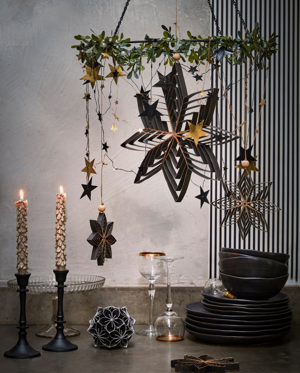 Zwarte metalen hanger, 40×46 cm (Øxh) € 9,99 omhuld met kunststof mistletoe guirlande € 3,99. Aan de mistletoe: diverse zwarte papieren ornamenten, per stuk vanaf € 2,49, goudkleurige metalen slinger met sterren, 210 cm (l) € 3,49 (ook in zilver). Op tafel: goudkleurige kaarsen met glitter, per 2 € 6,99, zwart papieren ornament bloem met glitter, 10 cm (Ø) € 5,99.