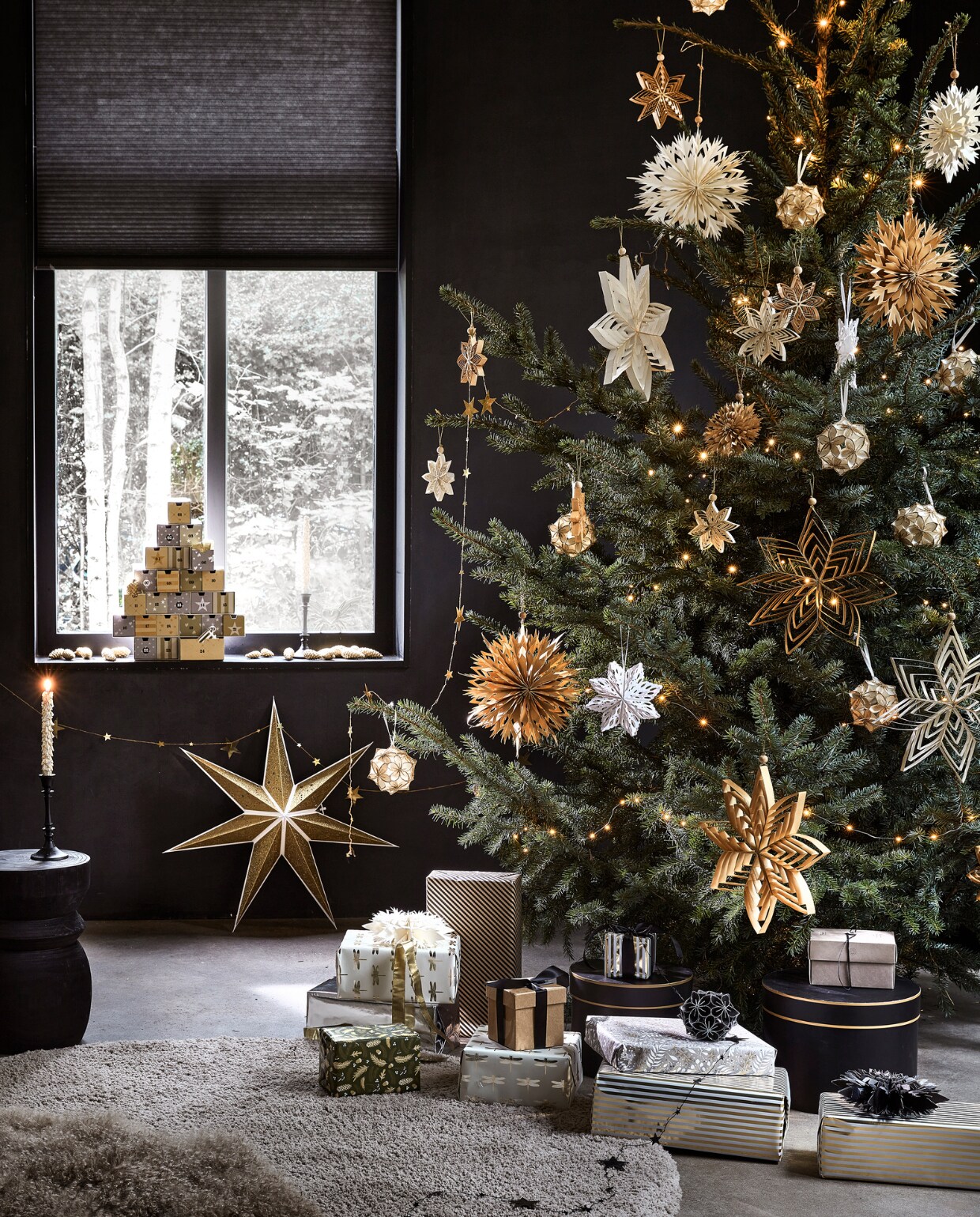 Kartonnen adventskalender € 27,99, goudkleurige kaars met glitter, per 2 € 6,99, goudkleurige papieren ster, 70×19 cm (Øxd) € 9,99, goudkleurige metalen slinger met sterren, 210 cm (l) € 3,49 (ook verkrijgbaar in zilver). In de boom: diverse papieren ornamenten in de kleuren goud, wit, zilver en zwart, per stuk vanaf € 2,49, zilverkleurige lichtslinger warm wit met 30 ledlampjes op batterijen inclusief display en timer, 300 cm (l) € 3,99.