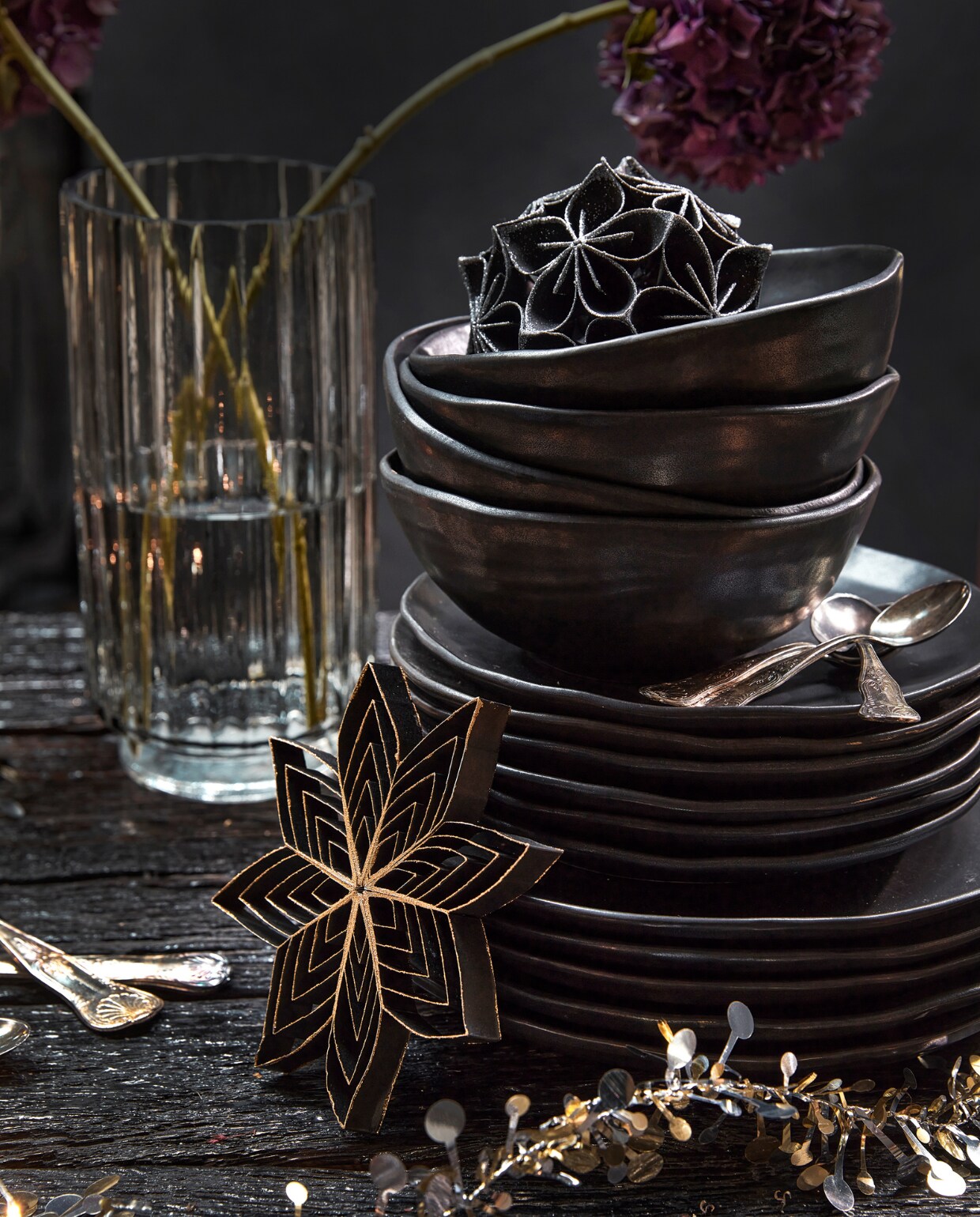 Zwart papieren ornament bloem met glitter, 10 cm (Ø) € 5,99, zwarte papieren ster van gerecycled papier, 15,5×3 cm (Øxh) € 2,69, goud- en zilverkleurige slinger met sterren en bolletjes van kunststof, 120 cm (l) € 7,99.
