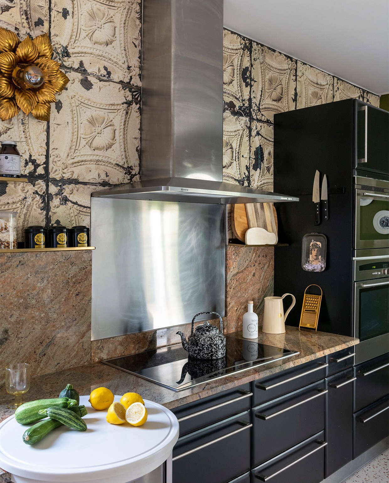 Keukenkastjes zwart geschilderd, vloer terrazzo-print linoleum van Gerflor, behang Merci voor Arte, muurverf kleur Bancha van Farrow&amp;Ball, tafel La Redoute, barkrukken vintage, hanglamp Ay Illuminate, planken House Doctor, muurlampen tweedehands, schilderij konijn Ibride, Chinese borden aan de muur antiek, gouden glazen Miss Étoile, theepot Merci Paris, waterkan Anthropologie. Overig onbekend of vintage.