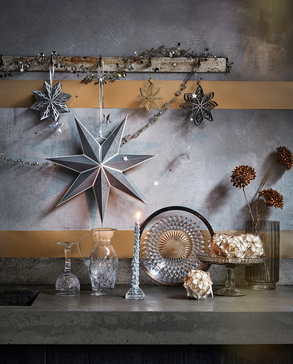 Zilverkleurige slinger met ijzeren belletjes, 120 cm (l) € 7,99, zilverkleurige metalen slinger met sterren, 210 cm (l) € 3,49, zilverkleurig papieren ornament, 15×1,5 (Øxd) € 2,99, zilverkleurige papieren ster, 70×19 cm (Øxd) € 6,99, goudkleurig ornament van gerecycled papier, 15,5×3 cm (Øxd) € 2,69, zwart ornament van gerecycled papier, 15,5×3 cm (Øxd) € 2,69, zilverkleurige kaars met glitter, per 2 € 6,99, wit papieren ornament bloem met glitter, 10 cm (Ø) € 5,99 (ook verkrijgbaar in zwart met zilver).
