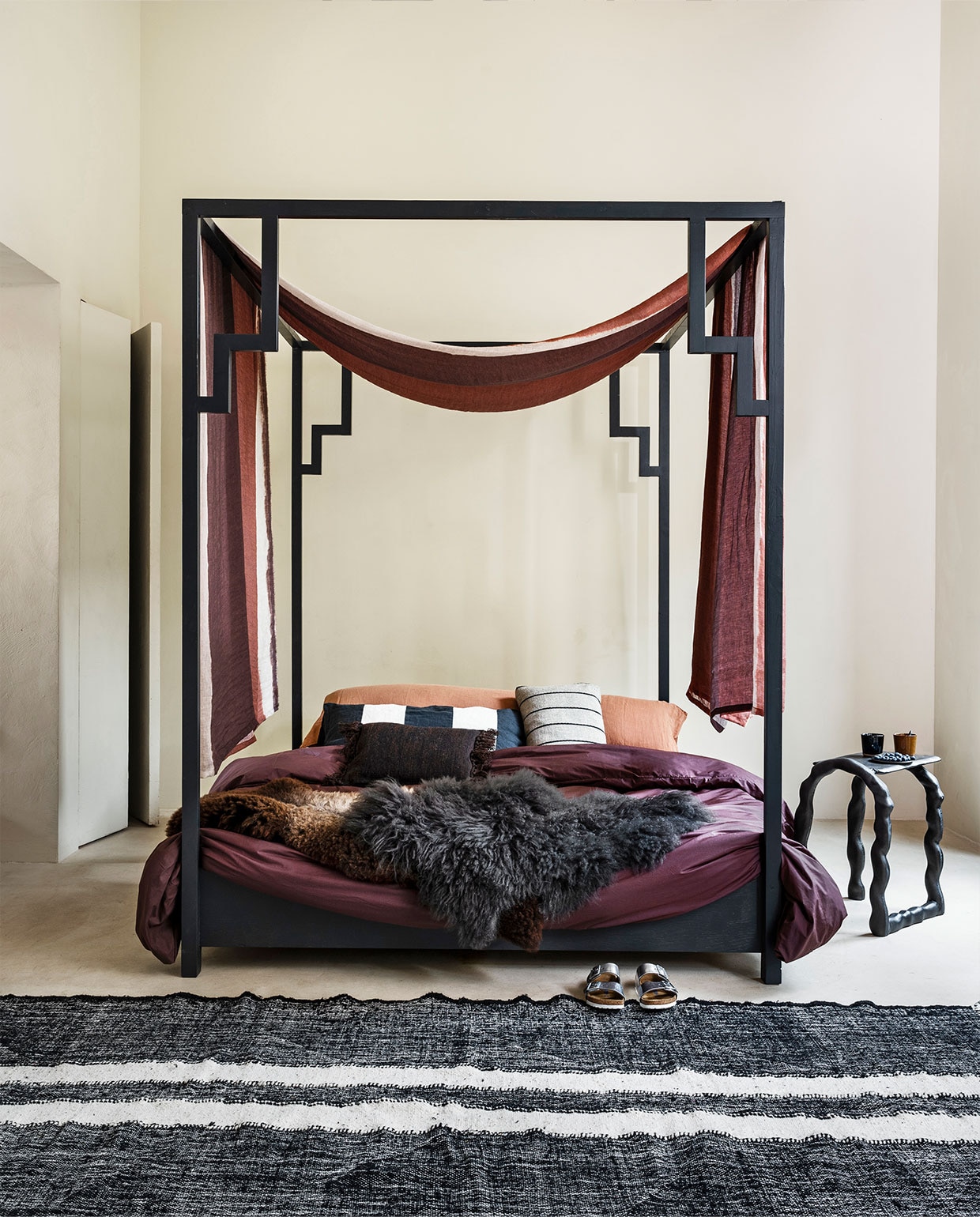 Hemelbed Oriënt, ontwerp Jeanette Spaansen, prijs op aanvraag (com-anders.nl). Linnen hemelstof Kanoko van Osborne &amp; Little, 306 cm breed, per meter € 126,- (wva.nl). Schapenvachten vanaf € 100,- (vanburenbolsward.nl). Stalen bijzettafel van Ferm Living, 45x32x54 cm (bxdxh) € 505,-, gestreept kussen van Ferm Living € 55,-, bruin kussen van Ferm Living € 55,- (misterdesign.nl). Mokken van Serax, per stuk € 10,50 (vtwonen.nl). Wollen vloerkleed, 300×120 cm € 289,-, langwerpige zwart-witte kussenhoes € 69,90 (10dayslifestyle.nl). Katoenen dekbedovertrek € 79,95, extra lang hoofd­kussen Bedmate 160×50 cm € 89,95, linnen sloop terra € 34,95 (suite702.com). Schoenen (birkenstock.com).