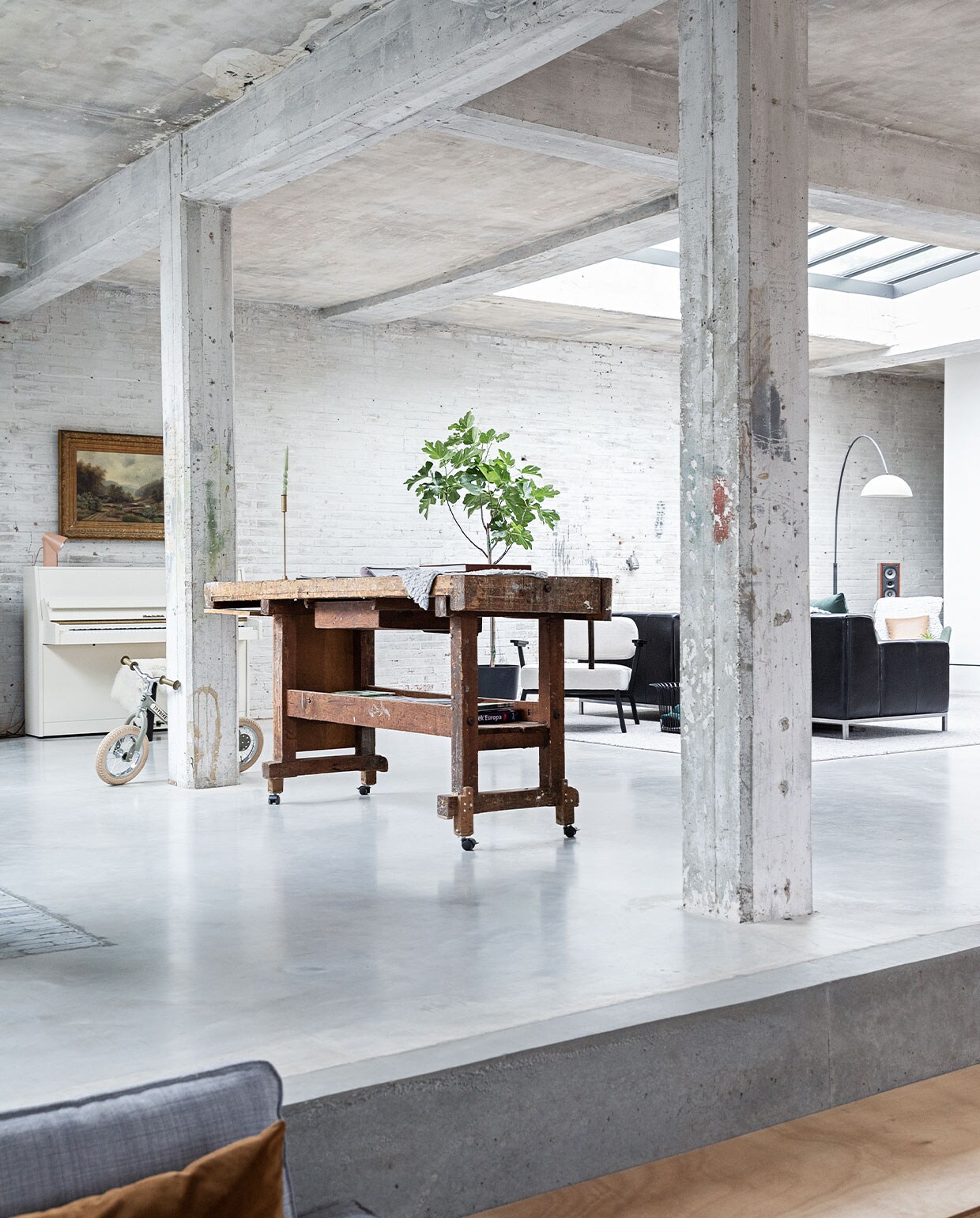 Vloer gevlinderd beton. Hardstenen waaltjes Spotted Bluestone. Werkbank erfstuk meubelfabriek. Kinderfiets Trybike. Witte fauteuil Ode Lounge Chair van Studio Henk.Bank Ikea. Booglamp Venled. Piano en schilderij erfstukken. Lamp op piano Elleke Rutten.