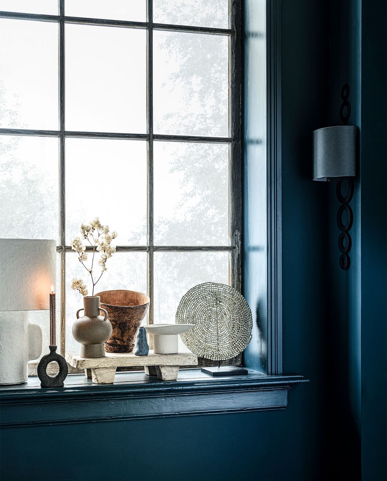 Op de wand krijtverf Steel Blue, per liter € 39,25 (pure-original.nl). Tafellamp € 219,- (loods5.nl/design). Zwarte keramieken kandelaar, 9,5x4x17 cm (lxbxh) € 97,-, houten dienblad op pootjes € 80,-, witte keramieken schaal op poot € 190,- (studio-yen.com). Beige vaas met oren € 12,99 (hm.com). Houten vaas € 22,90 (loods5). Blauwe minivaas € 14,95 (klevering). Object van schelpen op voet € 149,- (badenbaden). Wandlamp Selva van Vaughan € 905,-(wva).