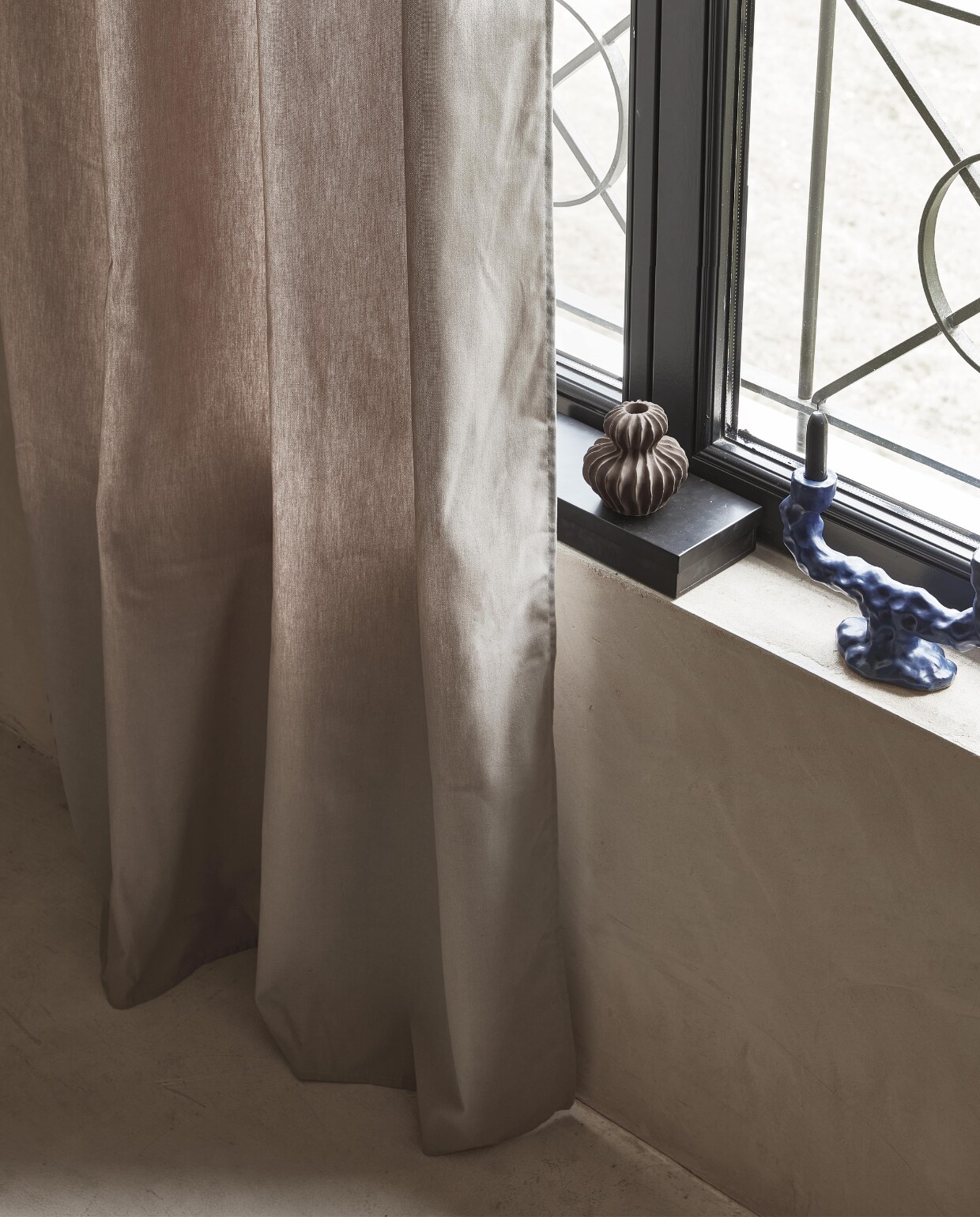 Taupe gordijn Denim Flax, lichtdoorlatend, met ring, 270×140 cm (hxb) € 55,99M.m.v. vtwonen.nl (blauwe kandelaar van Ferm Living), hm.com/home (bruin aardewerken vaasje), zarahome.com (zwart stalen kistje).
