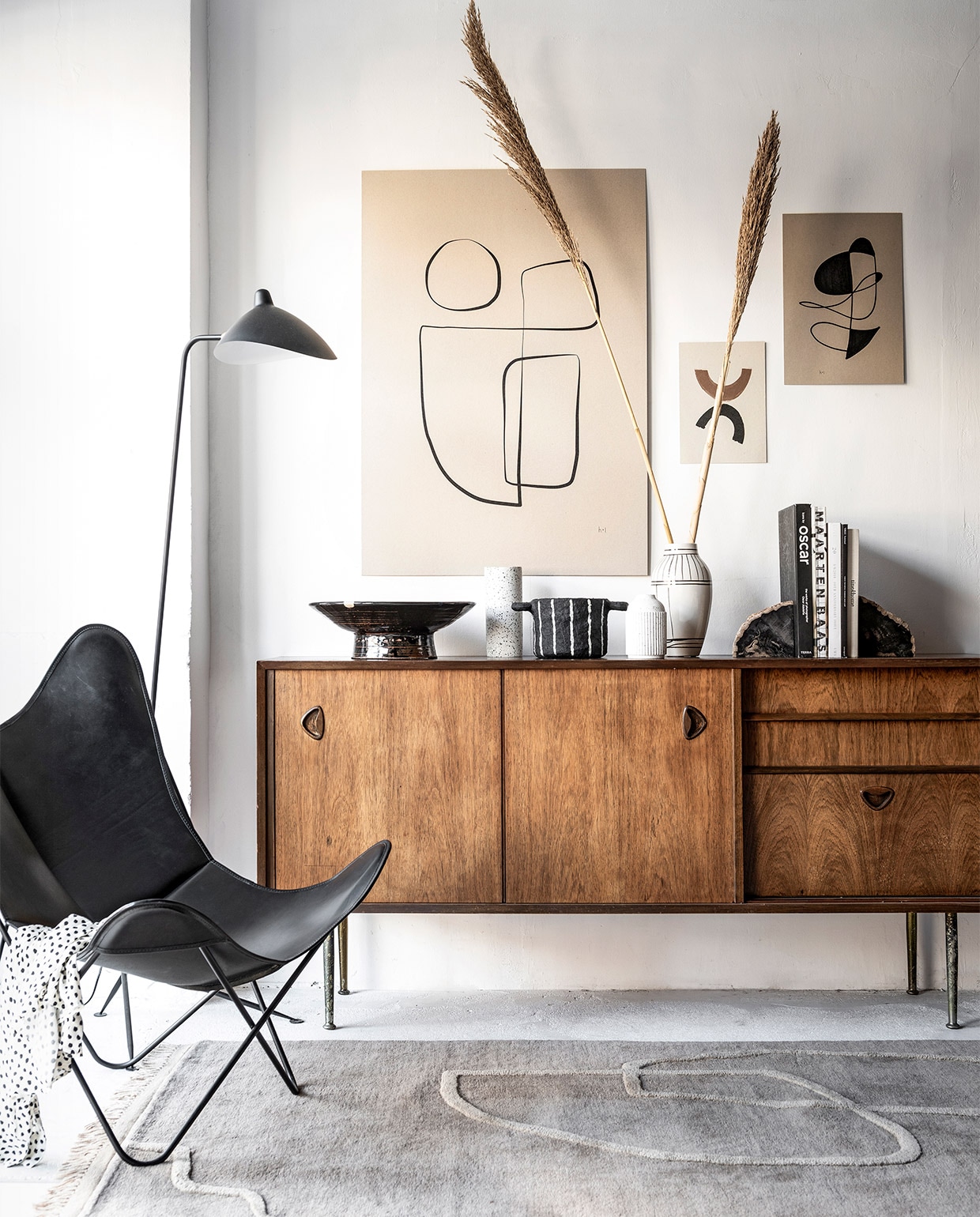 Abstracte inktlijntekening Sola van Henri &amp; Lydia € 6,79, abstracte inkttekening van Henri &amp; Lydia € 6,34, handgemaakte printable art van Miuus Studio set van 2 € 16,17 (etsy.com), alles geprint op grijsbord A4-formaat € 18,15, A3-formaat € 21,18, 70×100 cm € 53,85 (repros.nl). Schaal met lijnenspel € 475,-, lamp Replica Serge Mouille € 349,-, leren vlinderstoel voor Airborne € 999,-, teakhouten dressoir William Watting 19605 € 1295,- (cocooncollectables.nl). Terrazzo vaas van Zakkia € 39,- (restored.nl). Zwarte bloempot van papier € 33,50, wit keramiek vaasje met zwarte spikkels € 13,95 (storewithoutahome.nl). Witte vaas Outline € 26,33 (housedoctor.com). Handgemaakte boekensteunen van versteend hout € 149,- (studiosomee.com). Vloerkleed Desert Tufted Rug Sand, 140×200 cm € 599,- (fermliving.com). Boeken en blouse privébezit.