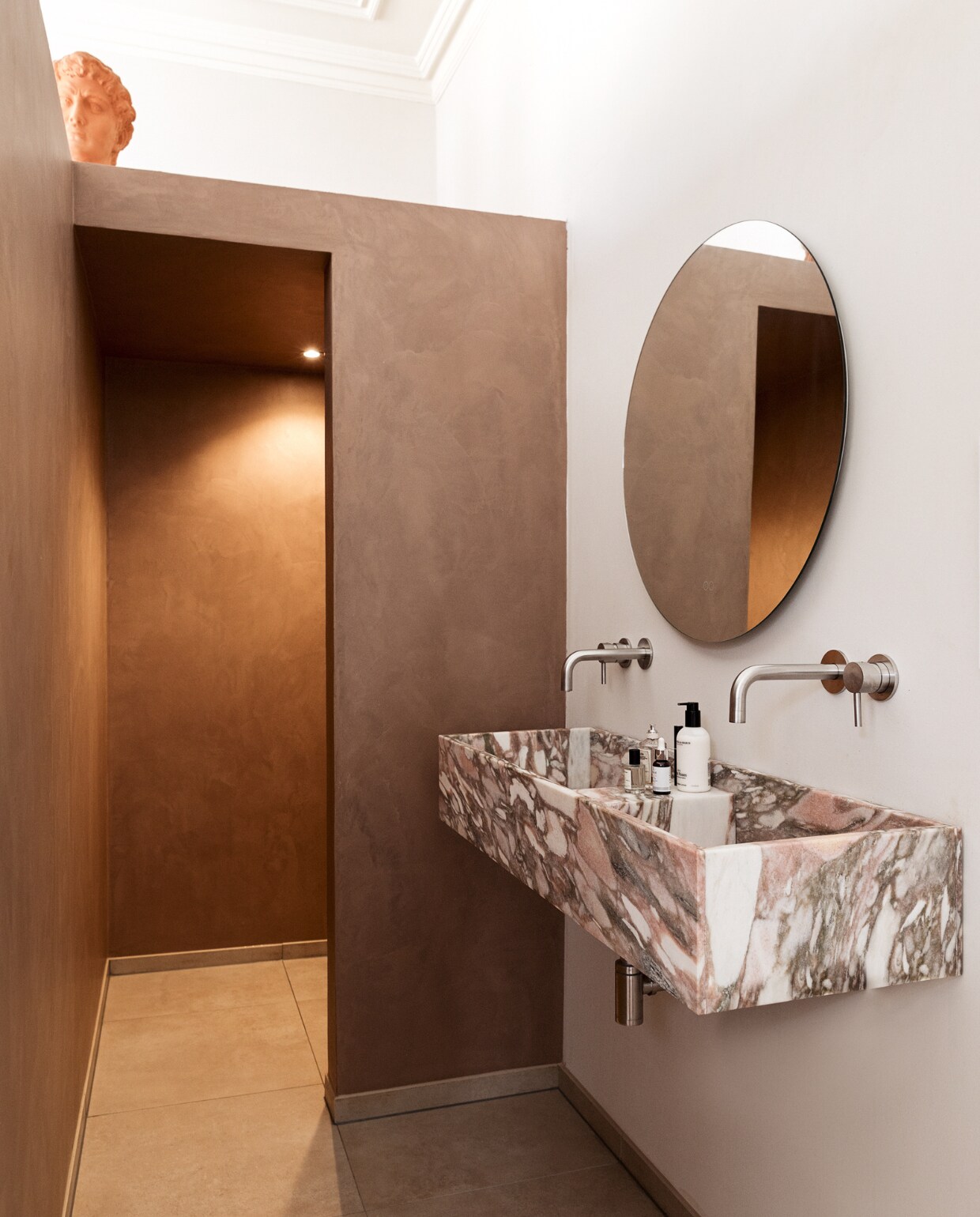 Ronde spiegel badkamerspiegels.nl. Wasbak heeft Louk nagetekend van een foto en laten maken door Swaal Natuursteen. Vloertegels Maxaro. Beton ciré in kleur Caramel Basebeton. RVS-kranen KraanDesign. Handzeep Marie-Stella-Maris. Gezichtsreiniging Image Skin Care.