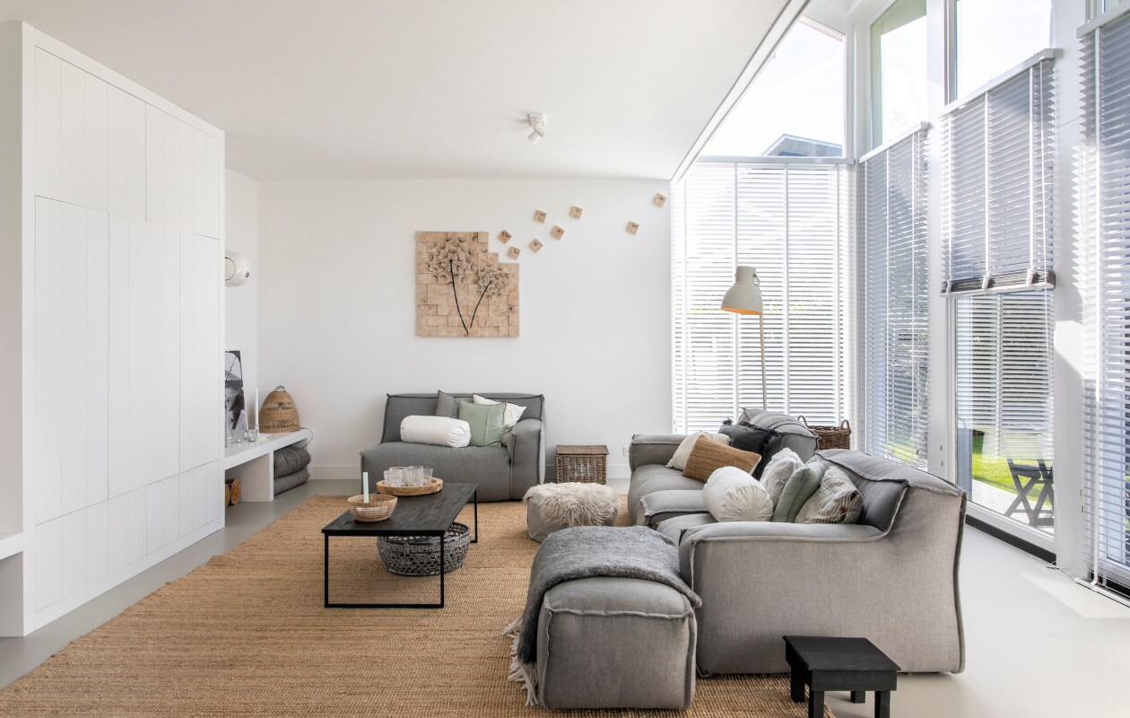 Bank De Troubadour, kunstwerk Paula Akkerman, groen kussen Maisons du Monde. Wit rolkussen, staande lamp, karpet Lohals van Ikea. Gestreept en donkergrijs kussen H&amp;M Home, naturel kussen en ronde manden Loods 5. Plafondlampen Rietveld Licht, raambekleding Joe Benelli, salontafel Ptmd, vacht Kwantum, lage vierkante bijzettafel Cooperz Wonen, plaid ’t Kethelhuys, vloer in kleur Nevada van Solo Gietvloeren, muurverf Fresh Linnen van Flexa. Overig onbekend.