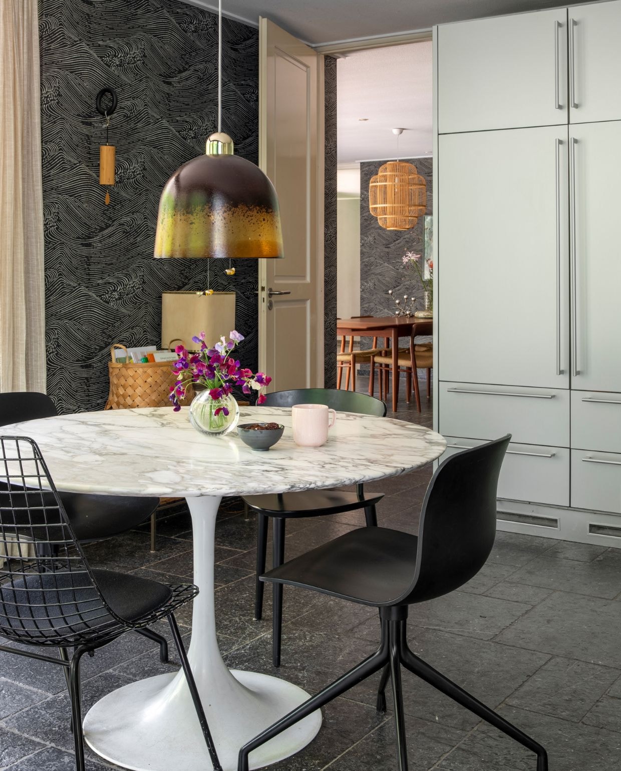 Behang Enso van Eijffinger. Marmeren vintage eettafel van Eero Saarinen, stoelen Hay en vintage, vaasje op tafel A.D. Copier van Royal Leerdam, kommetje en mok Iittala. Lamp boven tafel vintage, designer onbekend. Wandhaak Hay met Koshi gong Terra, gevlochten mand Søstrene Grene, tafellamp naast mand vintage, designer onbekend. Keuken Bulthaup, van vorige bewoners. Houten eettafel, stoelen en krukjes vintage Niels Otto Møller. Bamboe hanglamp Fine Asianliving. Vaas op houten tafel vintage, designer onbekend. Kandelaar Twiggy Squirrels via Pols Potten.