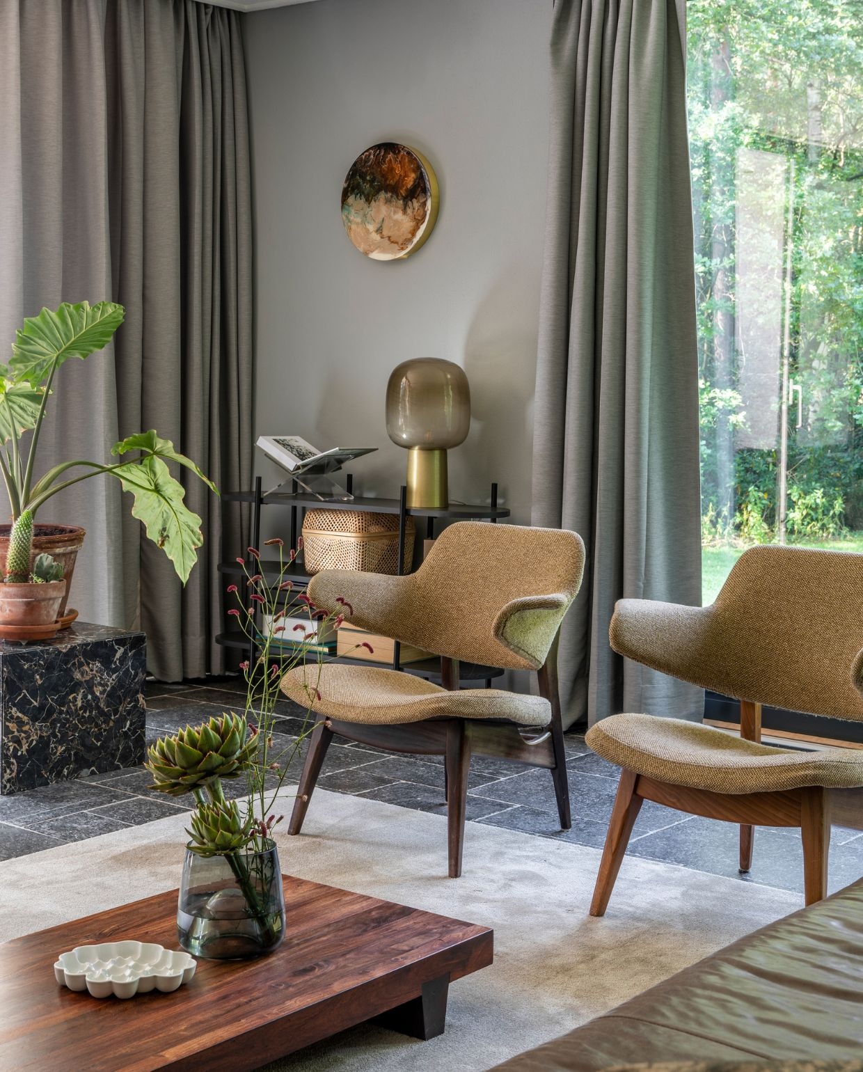 Muurverf Betongrijs Karwei. Gordijnen Ikea, vloer Belgisch hardsteen. Fauteuils vintage Louis van Teeffelen. Houten salontafel uit Japan via Catawiki, porseleinen schaal Bubble Dish van Studio Amber Isabel, glazen vaas op salontafel Loods 5. Vloerkleed Derby Beige, op maat laten maken bij Karwei. Marmeren kubus Marktplaats. Open kastje Leen Bakker, boekenstandaard Yessential, tafellamp House Doctor, opbergmand Ikea. Rond epoxykunstwerk Corine van Voorbergen. Bank Loods 5.