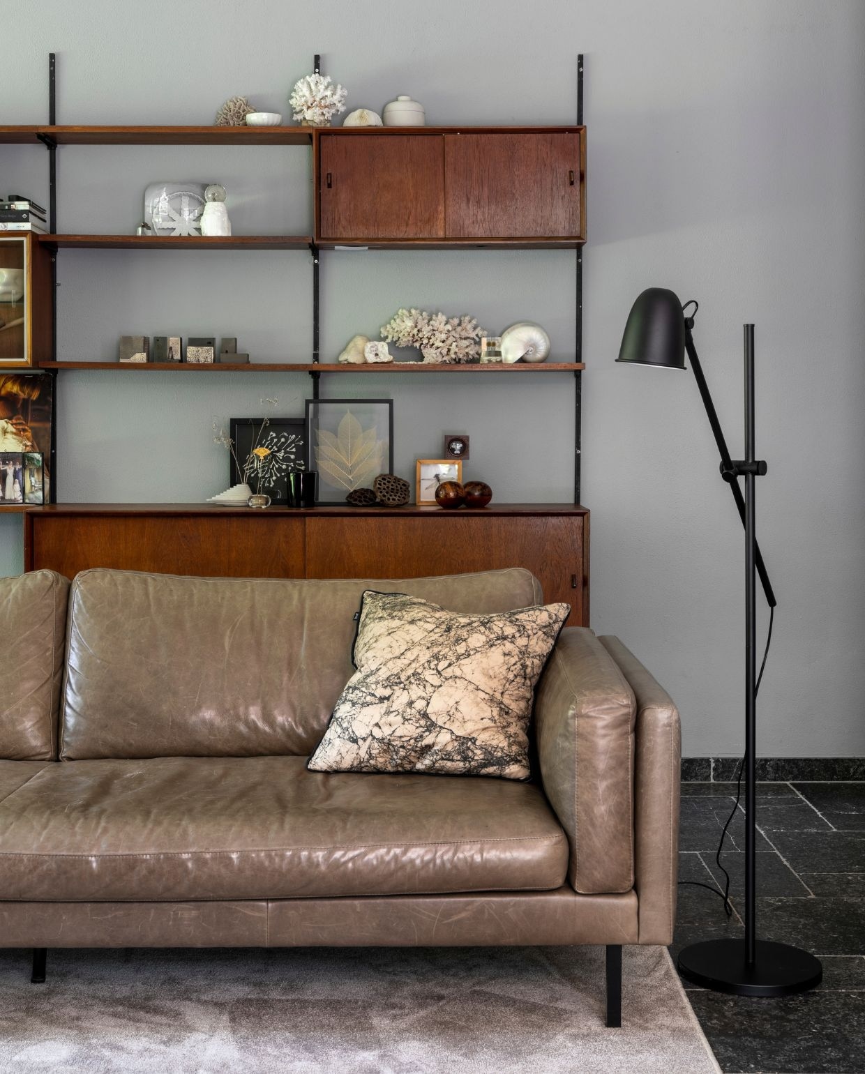 Muurverf Betongrijs Karwei. Wandkast vintage, designer onbekend. Bovenste plank: koraal vintage, wit schaaltje souvenir uit Japan, potje met deksel designer onbekend. Tweede plank: lens uit objectief voor porseleinen object van Studio Amber Isabel, stenen waxinelichthouder met paardebloem in epoxy. Derde plank: keramieken huisjes Studio Amber Isabel, koraal vintage met Nautilus parelmoerschelp. Onderste plank: betonnen vaasje en ikebana bloemhouder souvenirs uit Japan, ingelijste bloemen van porselein Studio Amber Isabel, gevonden libel ingelijst door opa, twee zaaddozen van lotussen uit Japan, zwarte tulpenvaas Pols Potten, koraal vintage, houten ronde bakjes souvenir uit Marrakech. Bank Loods 5, kussen op bank Ferm Living, zwarte lamp naast bank Ikea, vloerkleed Derby Beige Karwei.
