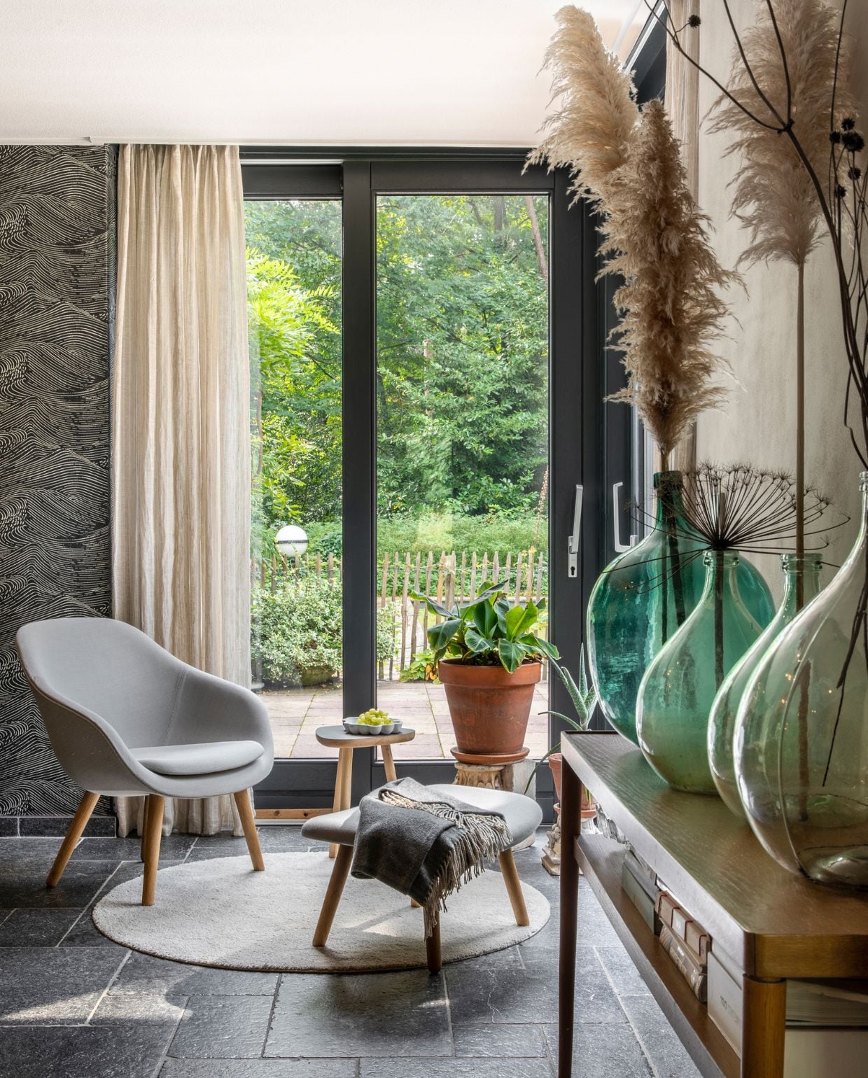 Behang Enso van Eijffinger, muurverf Elephants Breath Farrow &amp; Ball. Gordijnen Ikea. Rond vloerkleed Ikea, fauteuil met voetenbank Hay, plaid Loods 5. Bijzettafeltje designer onbekend, porseleinen schaal Bubble Dish van Studio Amber Isabel, antieke zuil met plant. Side table Ikea, gistingsflessen tweedehands, pluimen en droogbloemen uit eigen tuin.
