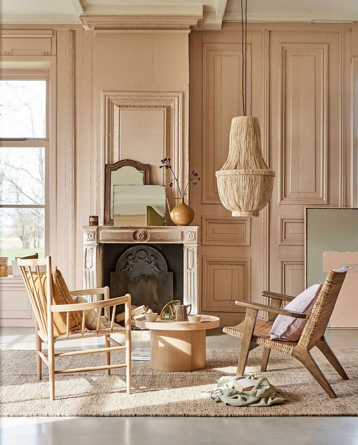 Roze tinten aan de muur met neutralen vloerkleed en schouw | Fauteuil links FD Møbler J82 Jørgen Baekmark Lounge Chai,, linnen tas, geruit kussen, geurkaars, mokken op tafel (letoile.store). Fauteuil rechts, vloerkleed, 200×300 cm, kunstwerk Abstract 100×120 cm, rechthoekige spiegel antieklook, vaas in mosterdkleur, glazen artwork op tafel (hkliving.nl). Zacht lila kussen van Bungalow, okergeel kussen, papieren dozen (scandicliving.nl). Oude spiegel met houten rand (com-anders.nl). Salontafel Plateau van Bolia, 34,3×61 cm (hxØ) (vtwonen.nl). Kroonluchter van gedroogde bananenbladeren (rivieramaison.com). Kleur op de wanden vergelijkbaar met Setting Plaster No.231 van Farrow &amp; Ball.