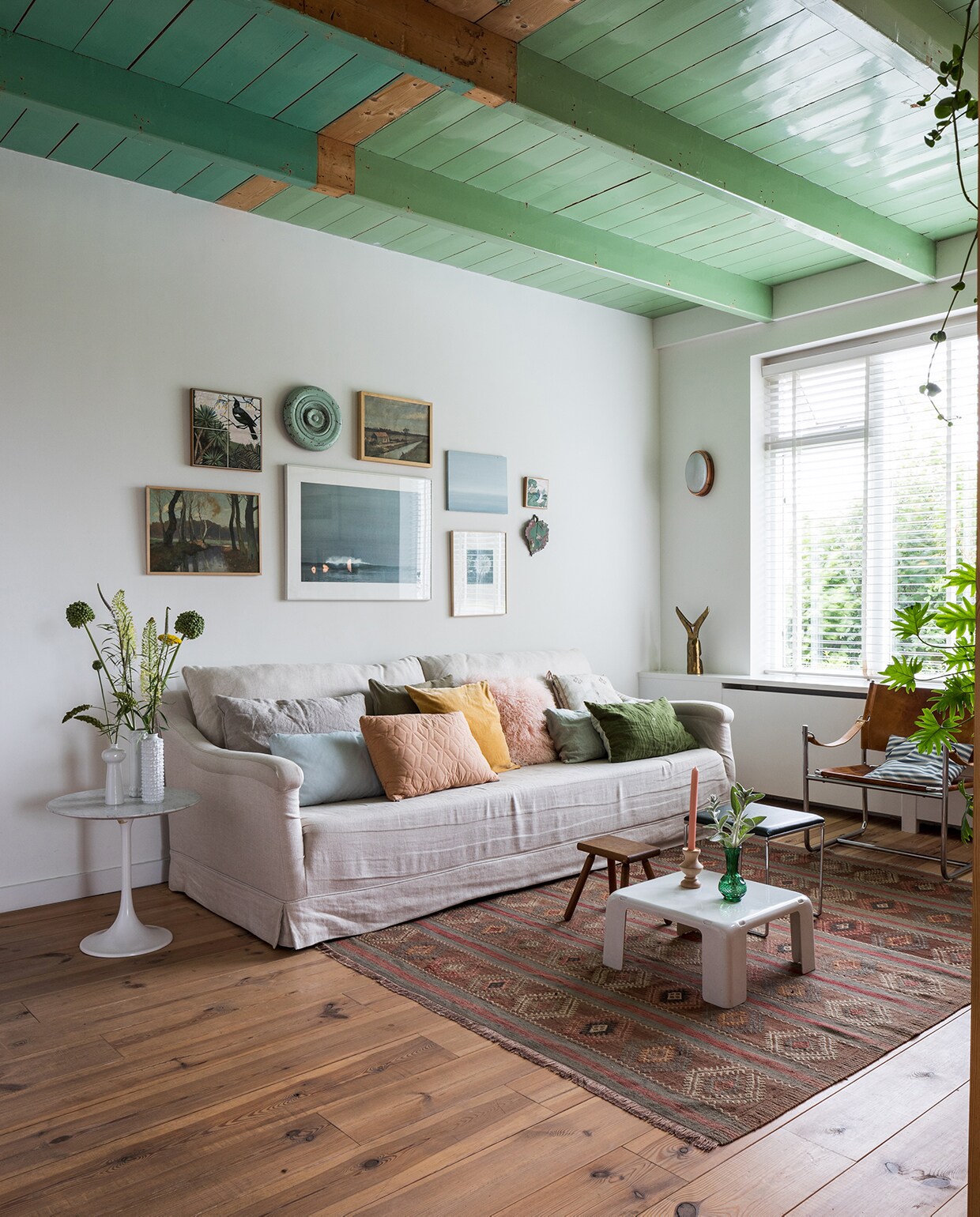 Bank Brechtje Olsthoorn Interiors, kussens zelfgemaakt, schapenvachtkussen Van Buren Bolsward. Groen linnen kussen, vazen, kandelaar, melkkruk, groene bijzettafel Sip &amp; Clara Pop-up Store. Tulp-tafel van Knoll erfstuk. Vintage kleed Richard Houpt Noordermarkt Amsterdam. Witte vintage bijzettafel B&amp;B Italia. Vintage buisframe stoel en wandlamp Marktplaats. Herkomst bronzen voorwerp onbekend. Aan de muur met de klok mee: tegel vogel uit Nieuw-Zeeland, groene deksel van ouderwets toilet, schilderij landschap rommelmarkt, horizonschilderij Auke van der Meulen, tegel uit Nieuw-Zeeland, aardewerken blad Trees In der Rieden, zeefdruk paardenbloem Ray Daal, grote foto cadeau gekregen, schilderij bomen kringloop.
