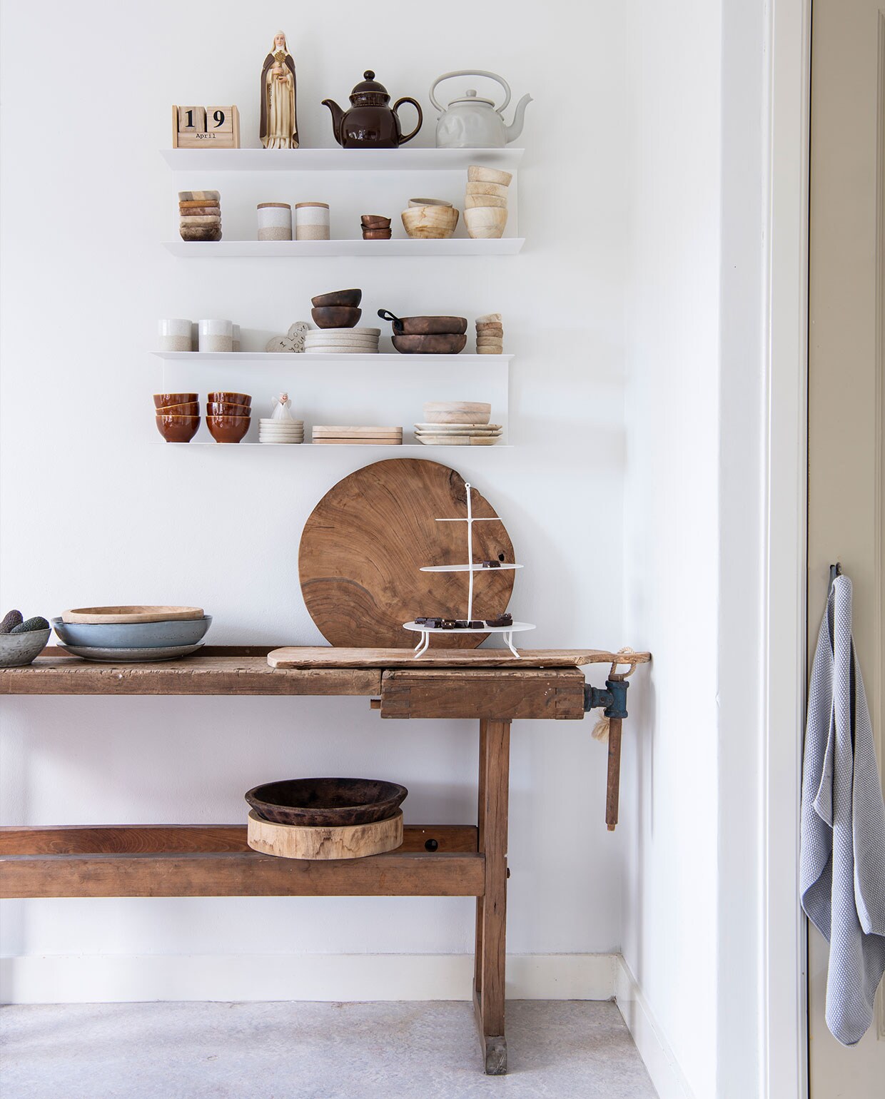 Wandplanken Ikea. Op de planken, van links naar rechts en boven naar beneden: kalender Twee21, beeld cadeau, bruine theepot Tablejoy, grijze theepot Kastanje Wonen, houten bakjes kringloopwinkel, voorraadpotten en aardewerken kommen Bloomingville, houten kommen Anne-Marie Wonen, kopjes en borden Bloomingville, houten kommen NADesign, geglazuurde kommen Tablejoy, bordjes Bloomingville, plankjes Anne-Marie Wonen, aardewerken borden en kom Bloomingville. Werkbank herkomst onbekend. Vijzel Passie Wonen Soest. Blauw-grijze schaal en grijs bord Broste Copenhagen via Twee21. Diverse houten kommen en snijplanken Anne-Marie Wonen. Etagère K’willeminhuis. Theedoek Södahl.